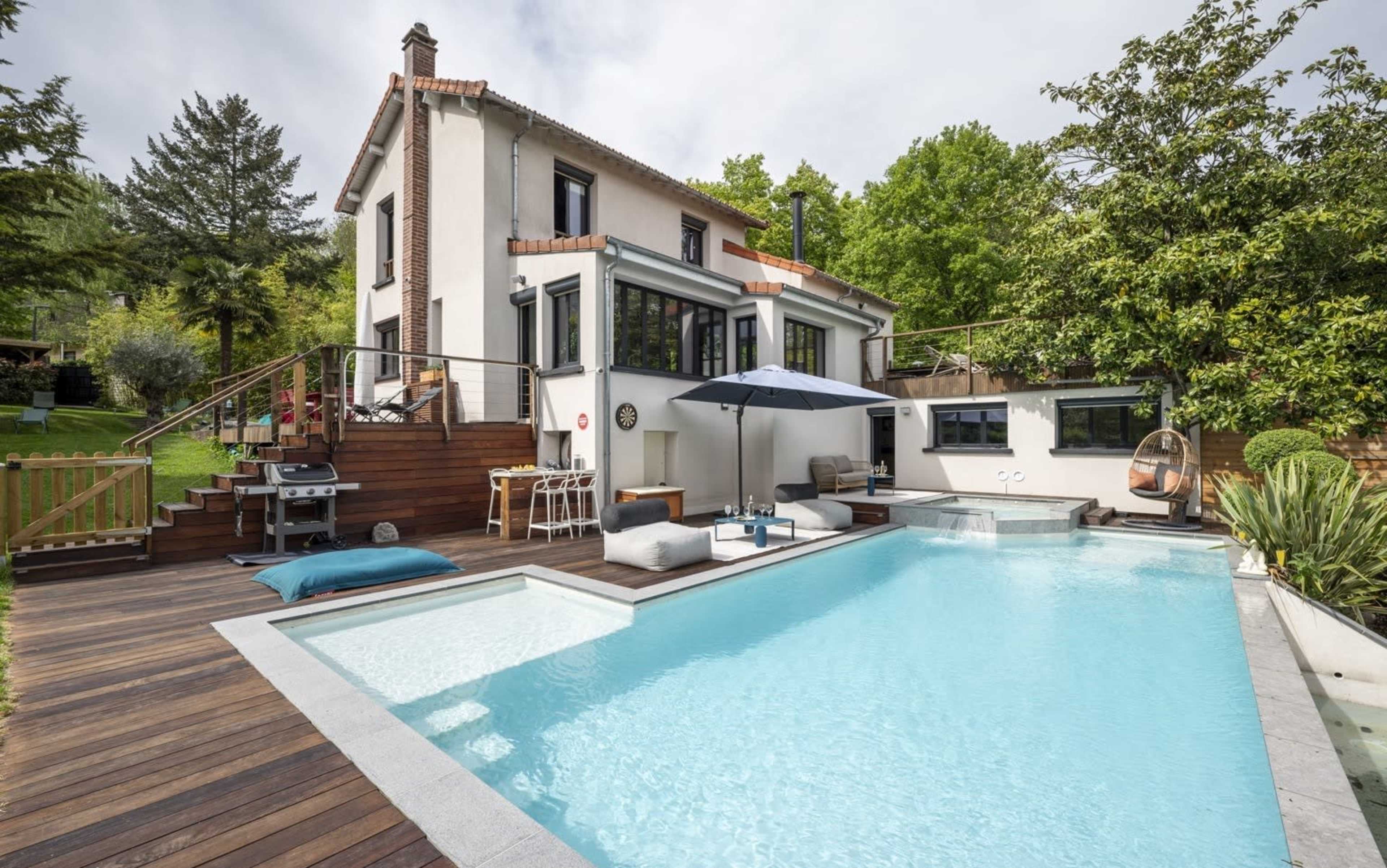 A modern house with a swimming pool and a patio area features outdoor seating, a grill, and lush greenery surrounding it.