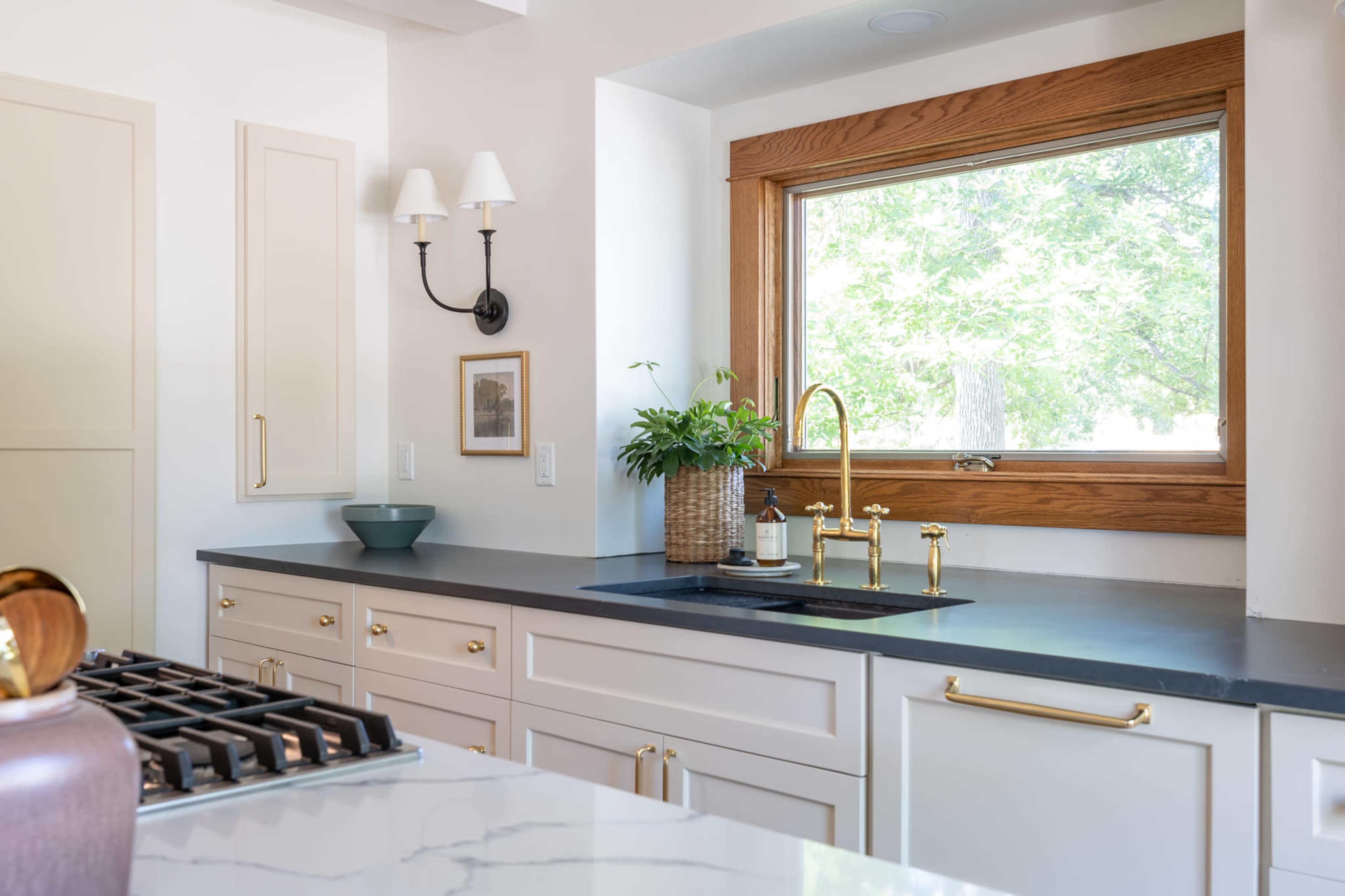A modern kitchen with light-colored cabinetry, a black countertop, and a window framed in wood, featuring a sink and a potted plant nearby.