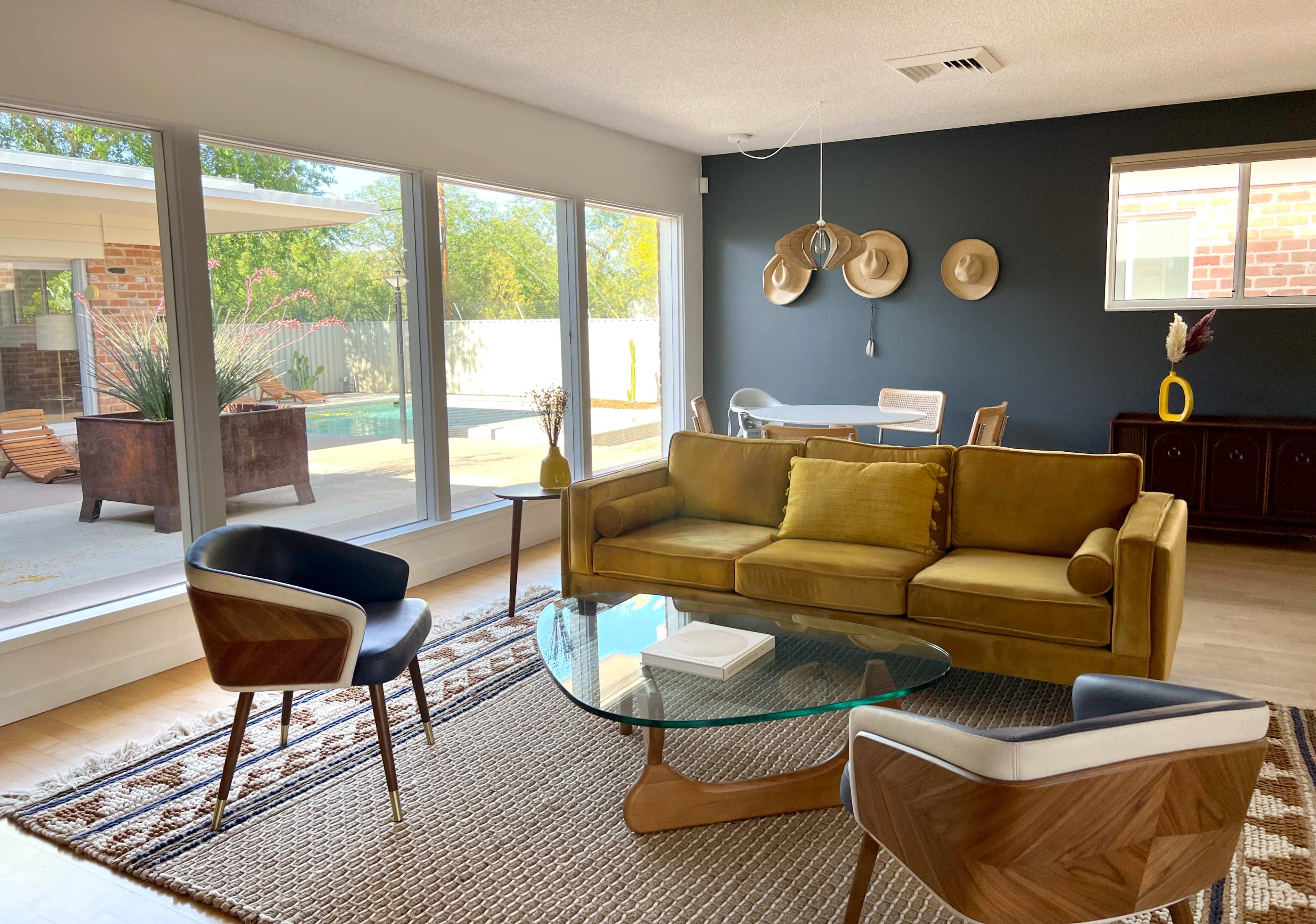 The living room features a yellow sofa, glass coffee table, and two modern chairs, with large windows overlooking a pool area.