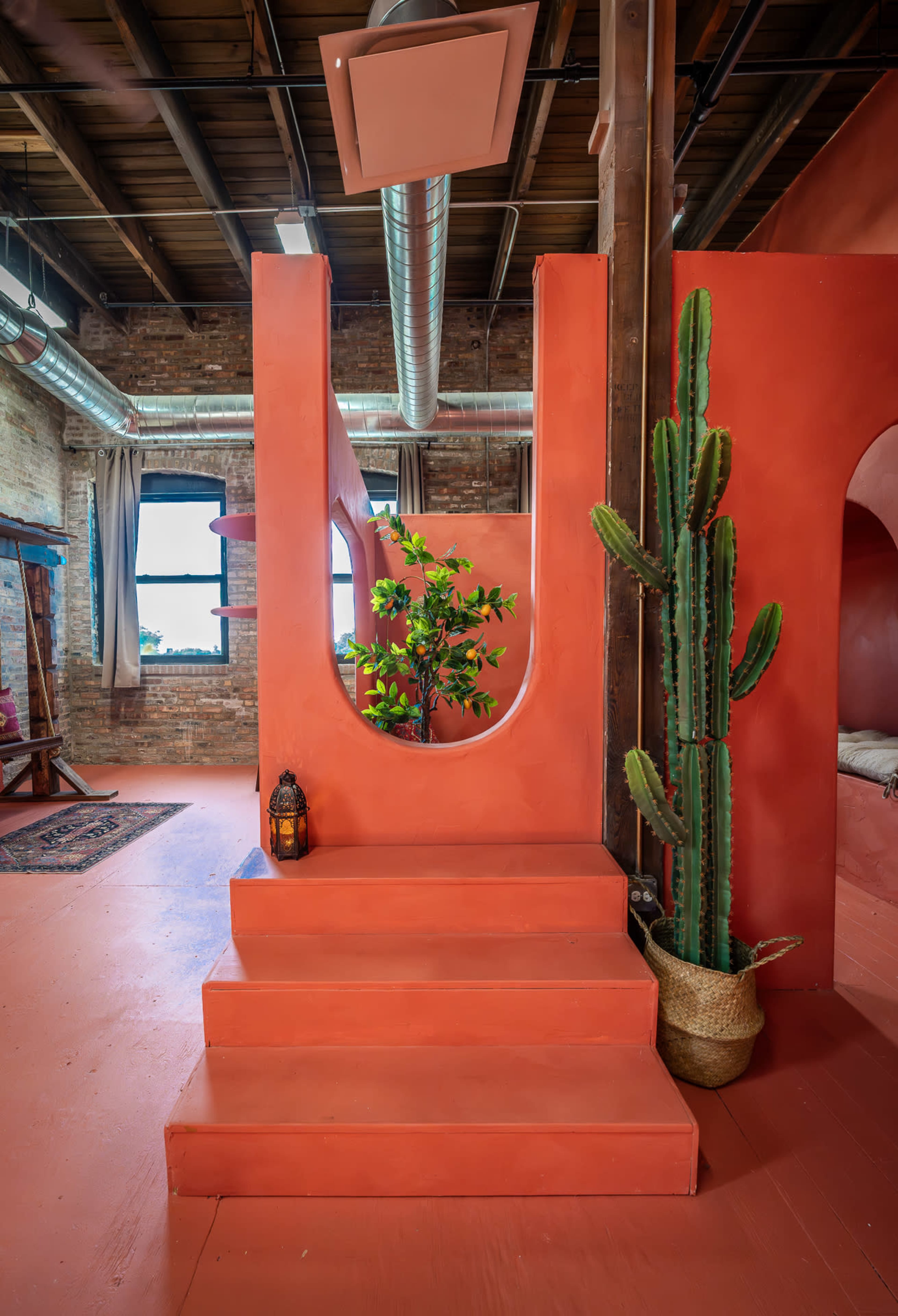 The image shows a vibrant interior space featuring orange walls, a staircase leading up, a potted cactus, and a small tree visible through a round cutout.