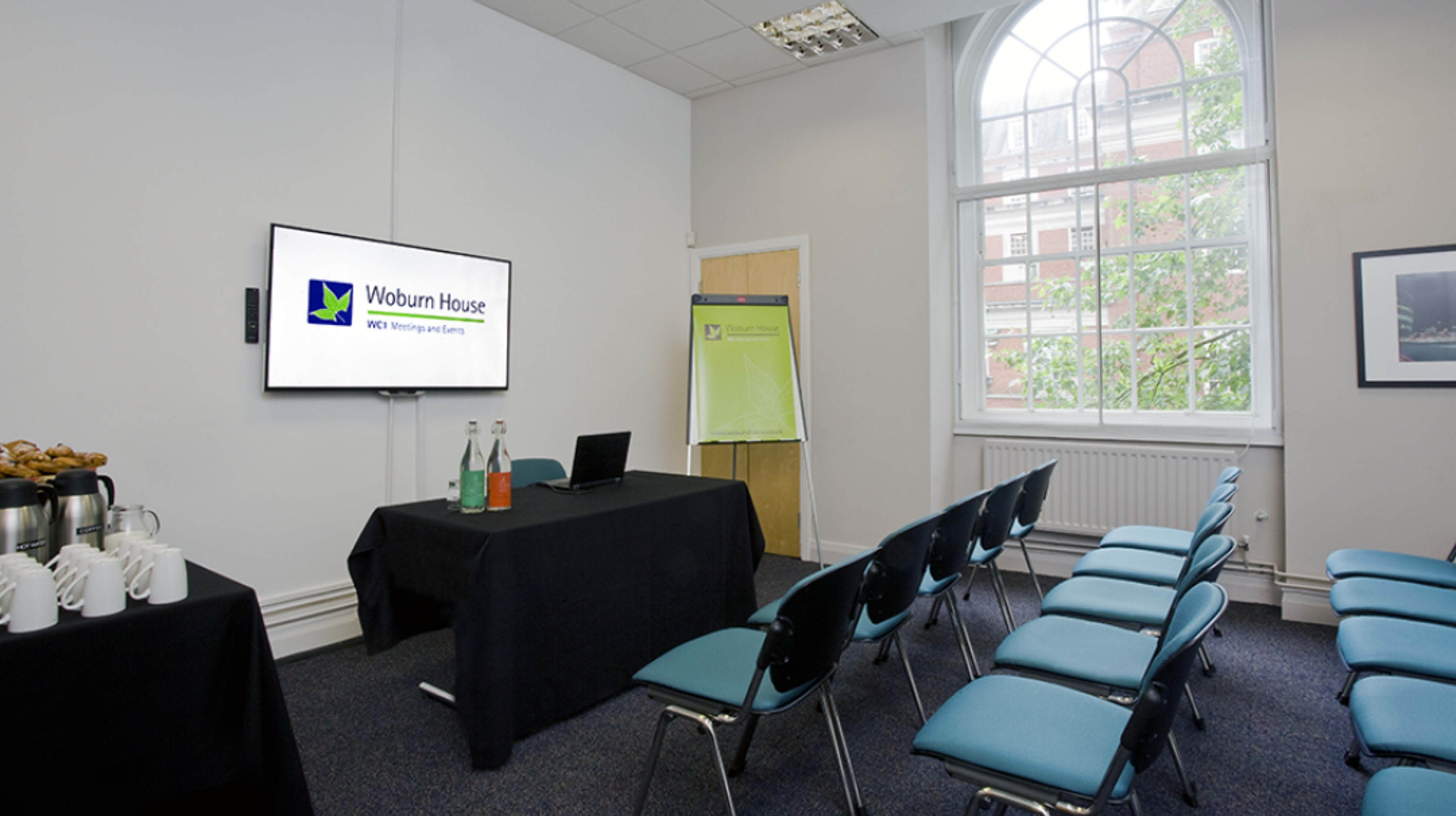A meeting room is set up with a monitor displaying a logo, a table with beverages, and several rows of chairs.