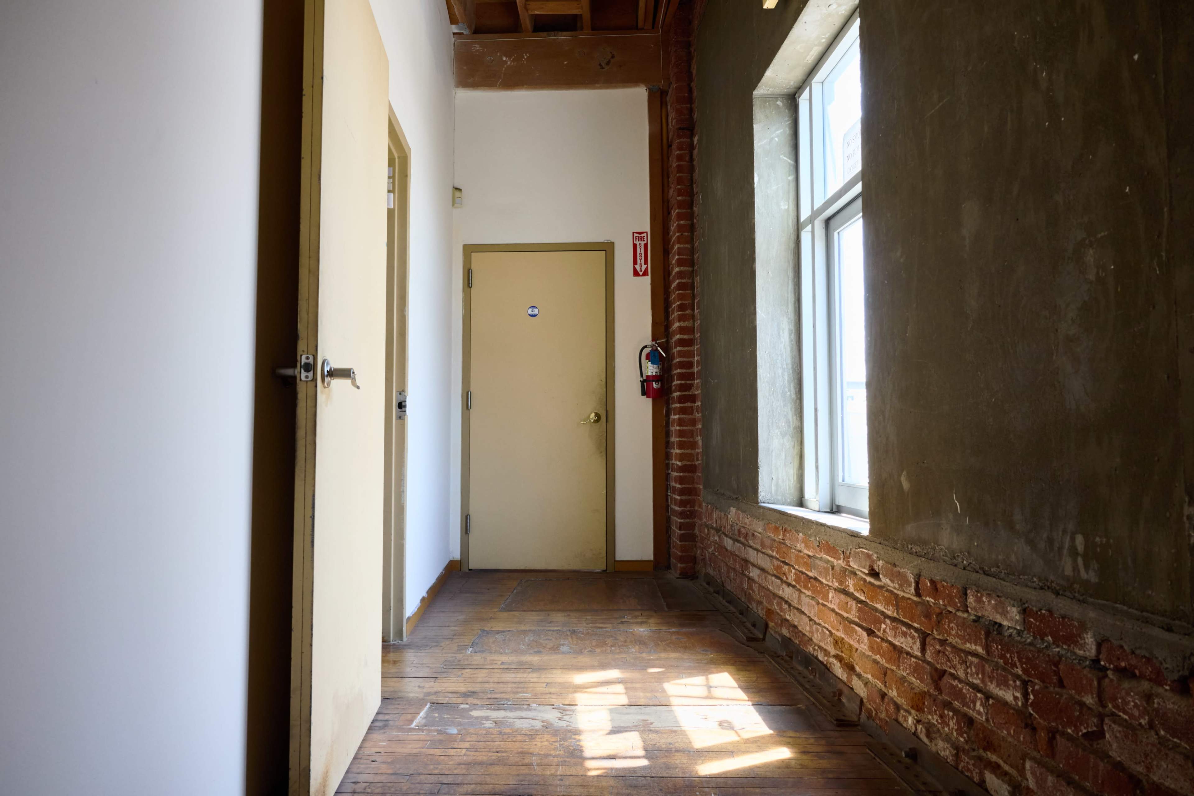 The image shows a narrow hallway with a textured wall on one side, two doors, and a window allowing natural light to enter.