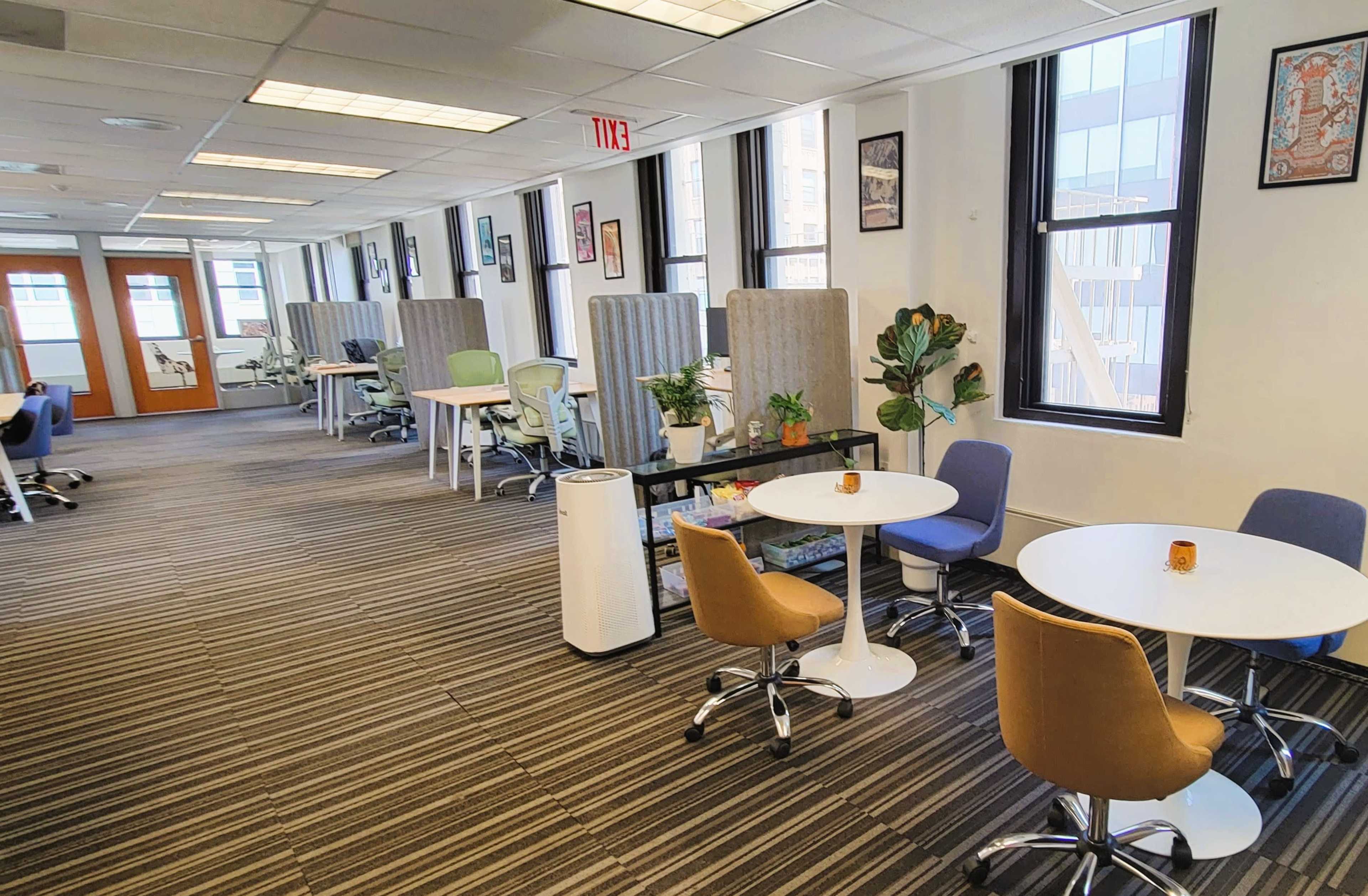 A bright, open office space with individual workstations separated by fabric partitions and various plants, along with round tables and chairs arranged throughout.