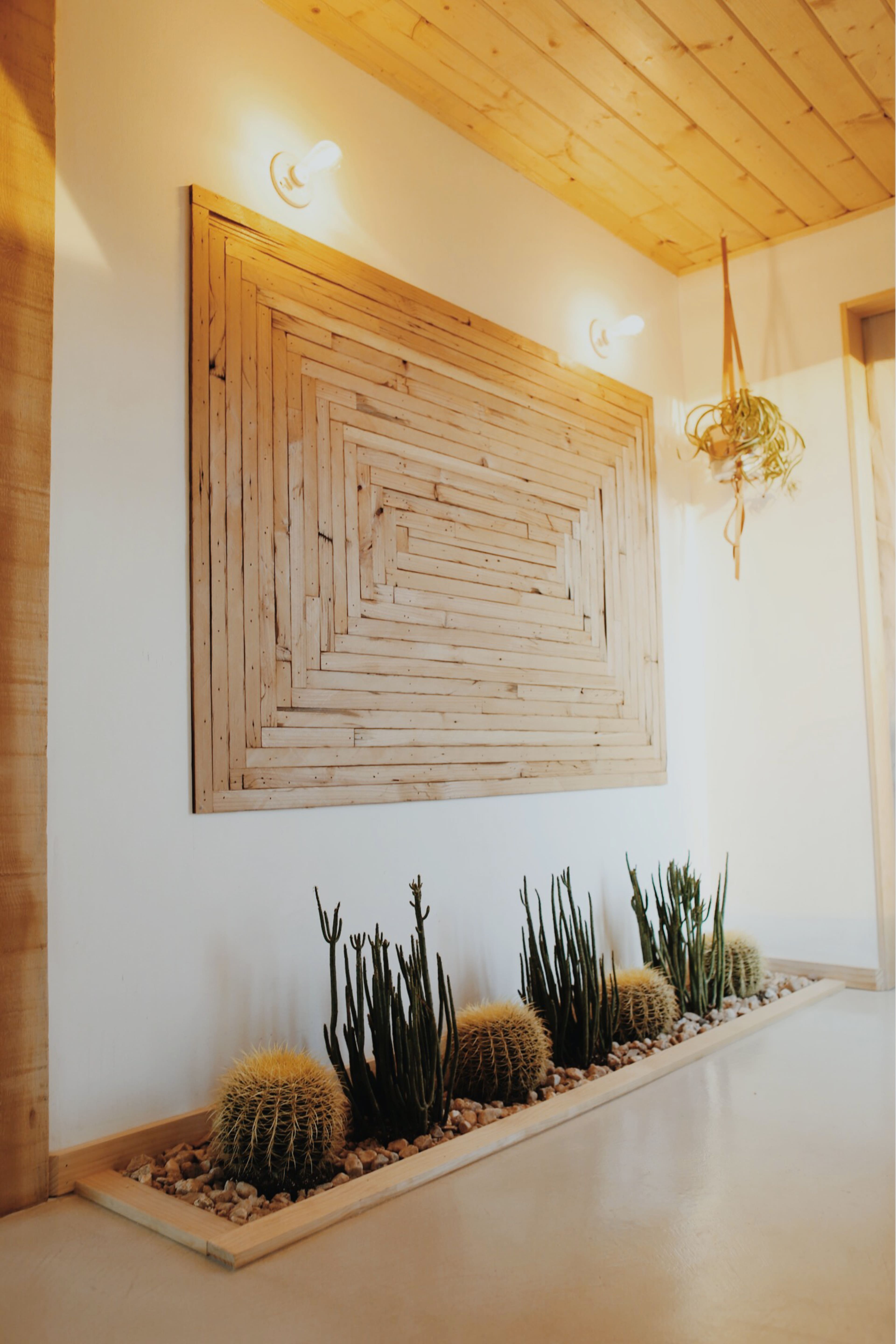 The image depicts a hallway featuring a wooden geometric wall art piece, with ornamental cacti and pebbles arranged beneath it.