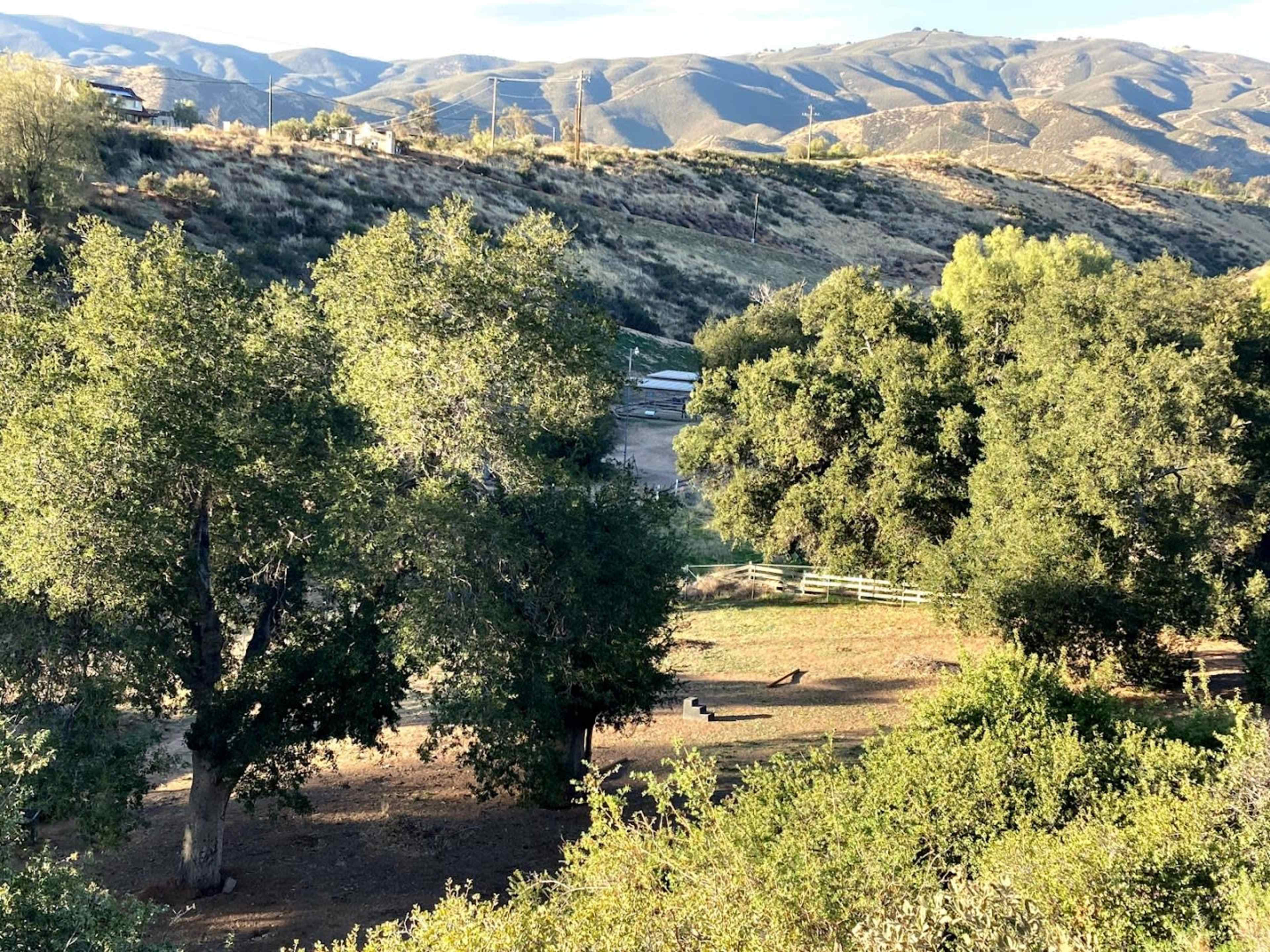 A mountainous landscape with green trees and a dirt area, partially bordered by a white fence.