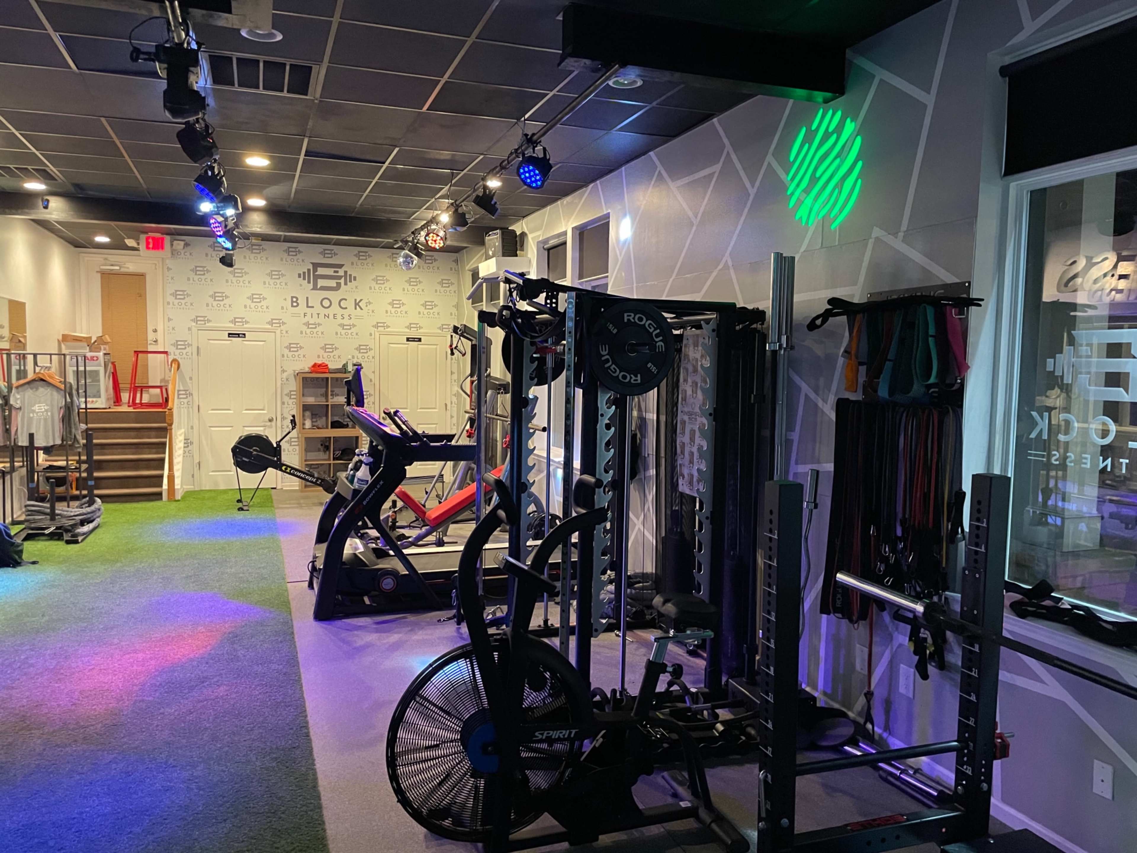 A fitness studio featuring exercise equipment, including a squat rack and stationary bikes, with colorful lighting and grass-like flooring.