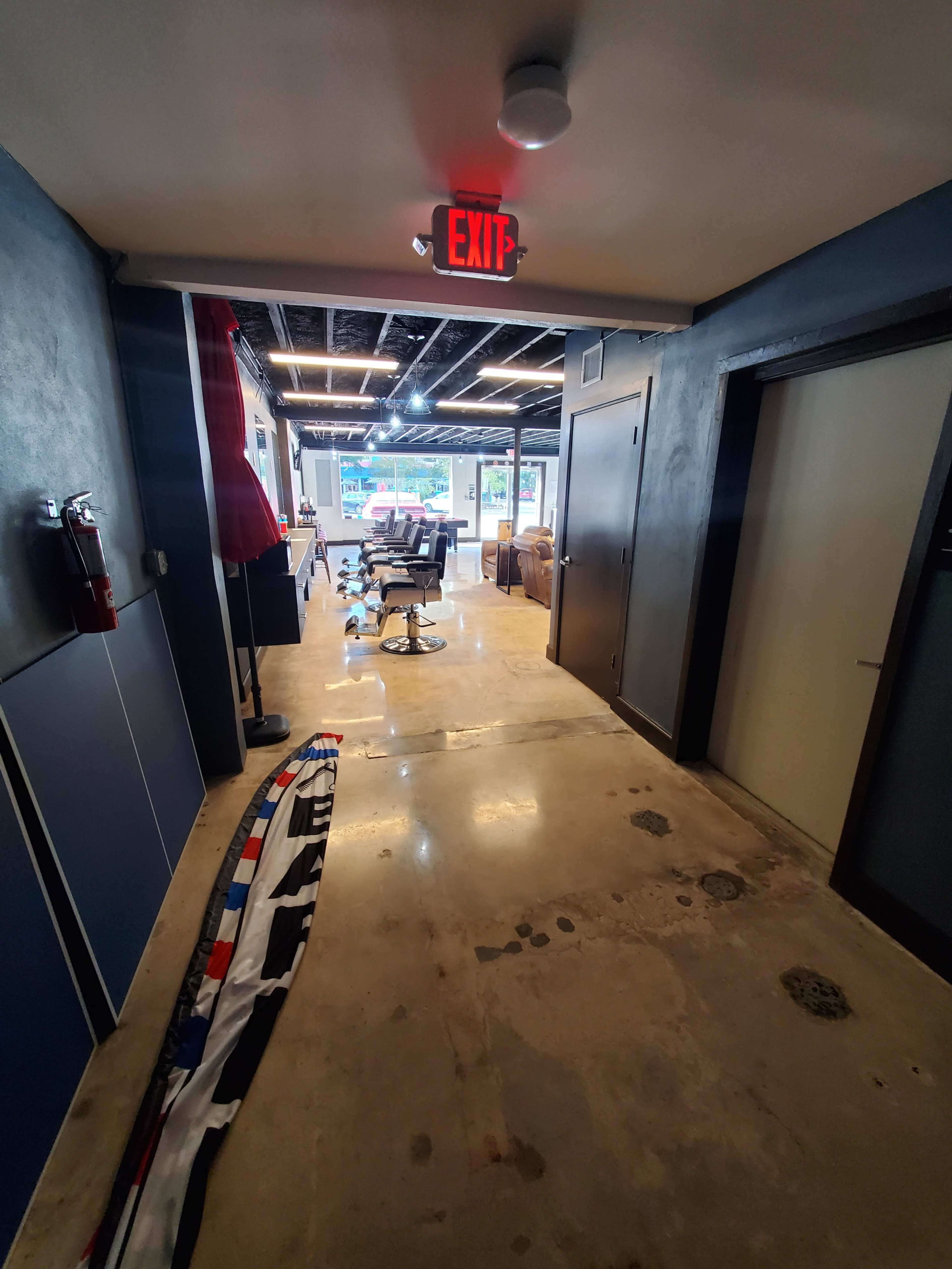 A long corridor with polished concrete flooring leads to a brightly lit area at the far end, featuring a barber chair and a seating area.