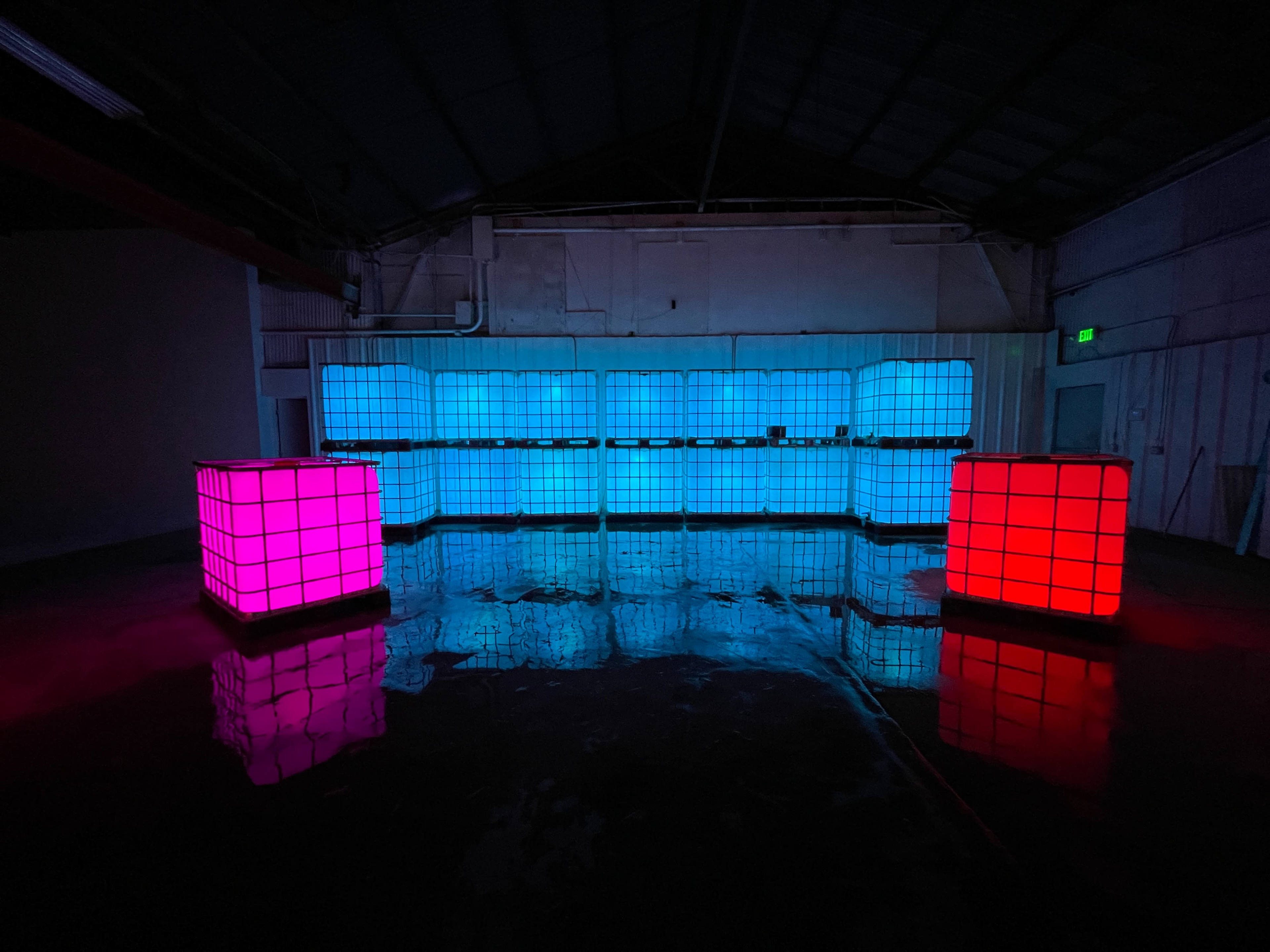 The image shows a dimly lit warehouse space with glowing colored cubes, one pink and the other blue, reflecting on the wet floor.