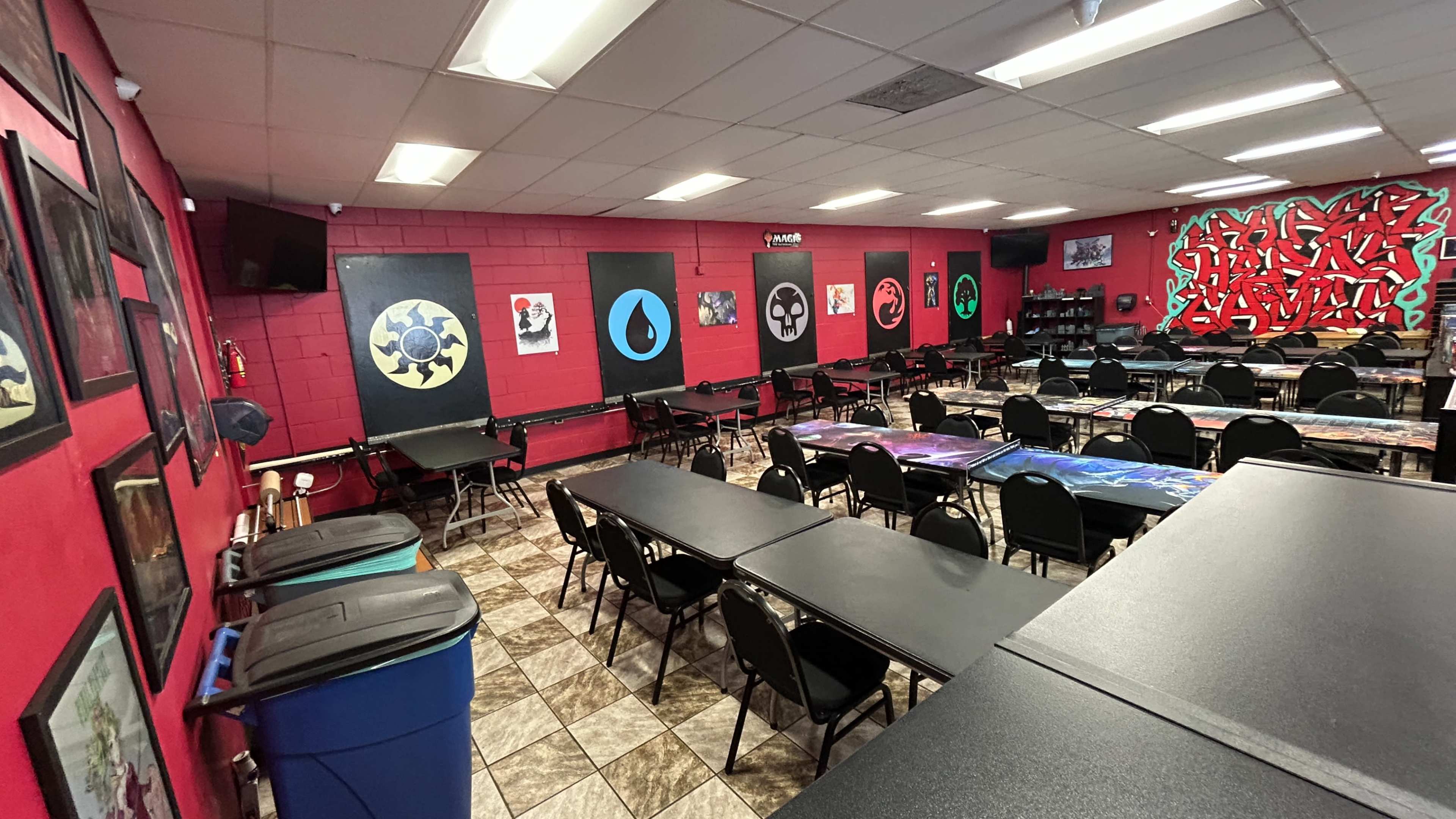 The image shows a spacious game room with rows of tables and chairs, decorated with vibrant artwork and gaming-themed posters on the walls.