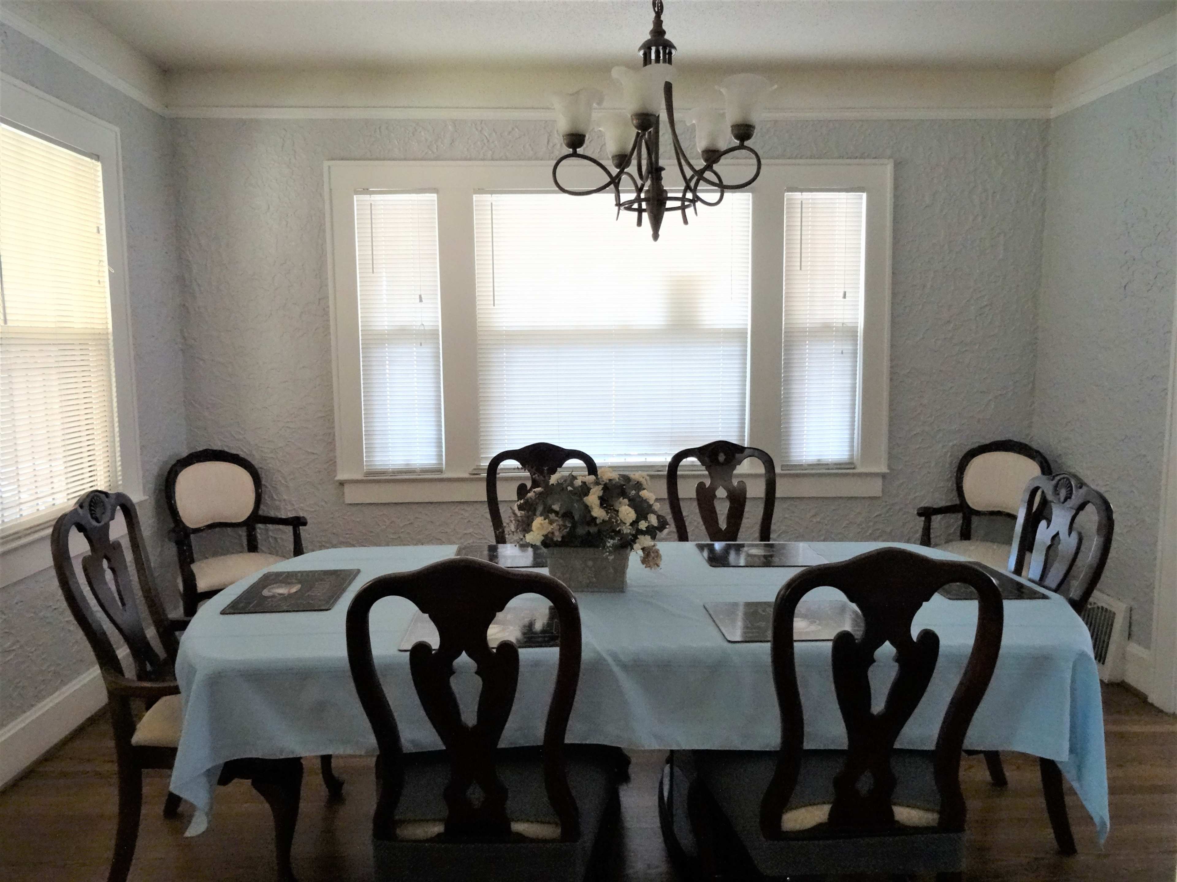 A dining room features a long table with a light blue tablecloth surrounded by six wooden chairs, with a floral centerpiece and a chandelier above.