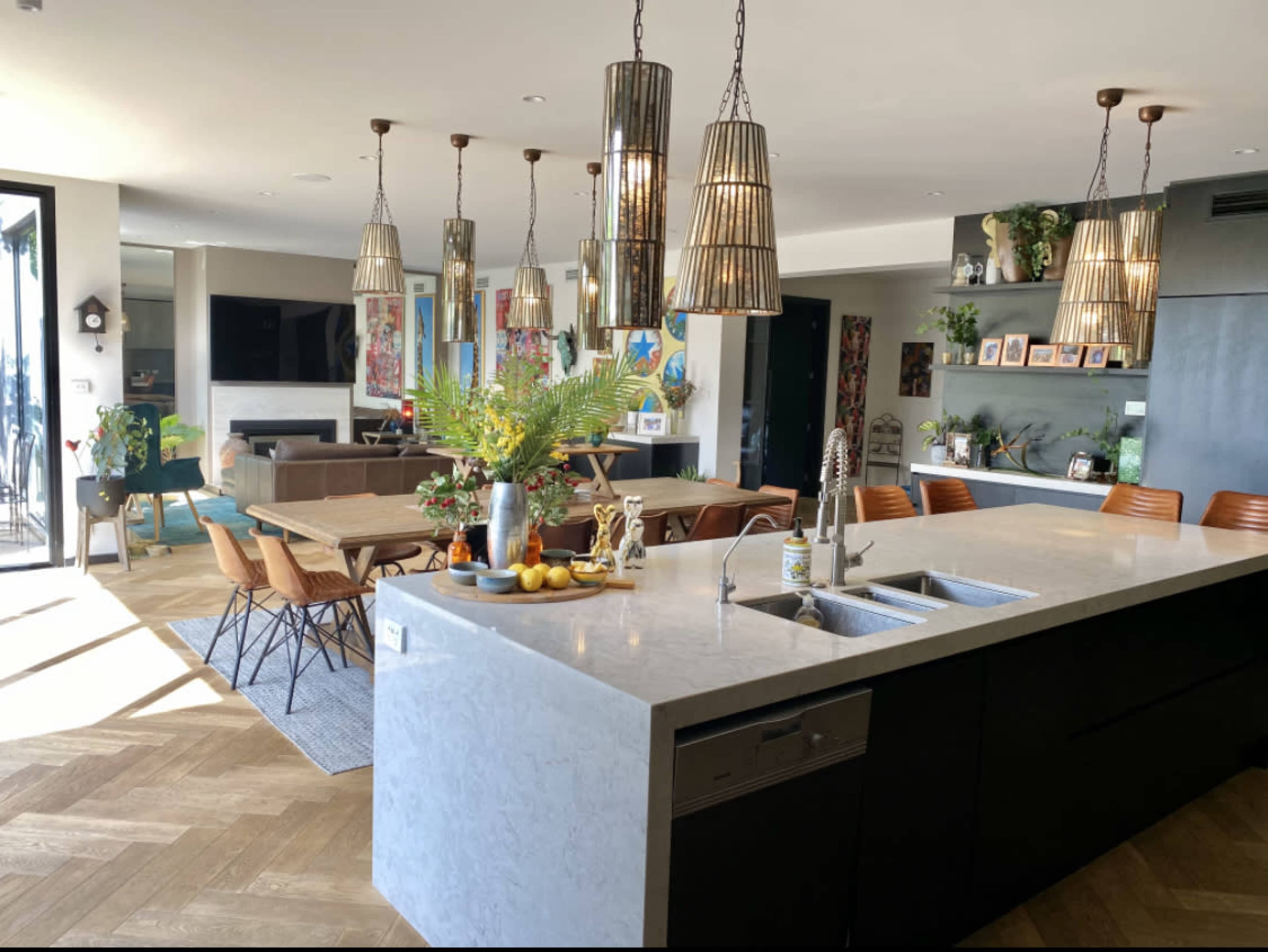 The image shows a modern kitchen with a large island, pendant lighting, and a dining area featuring a wooden table surrounded by chairs.