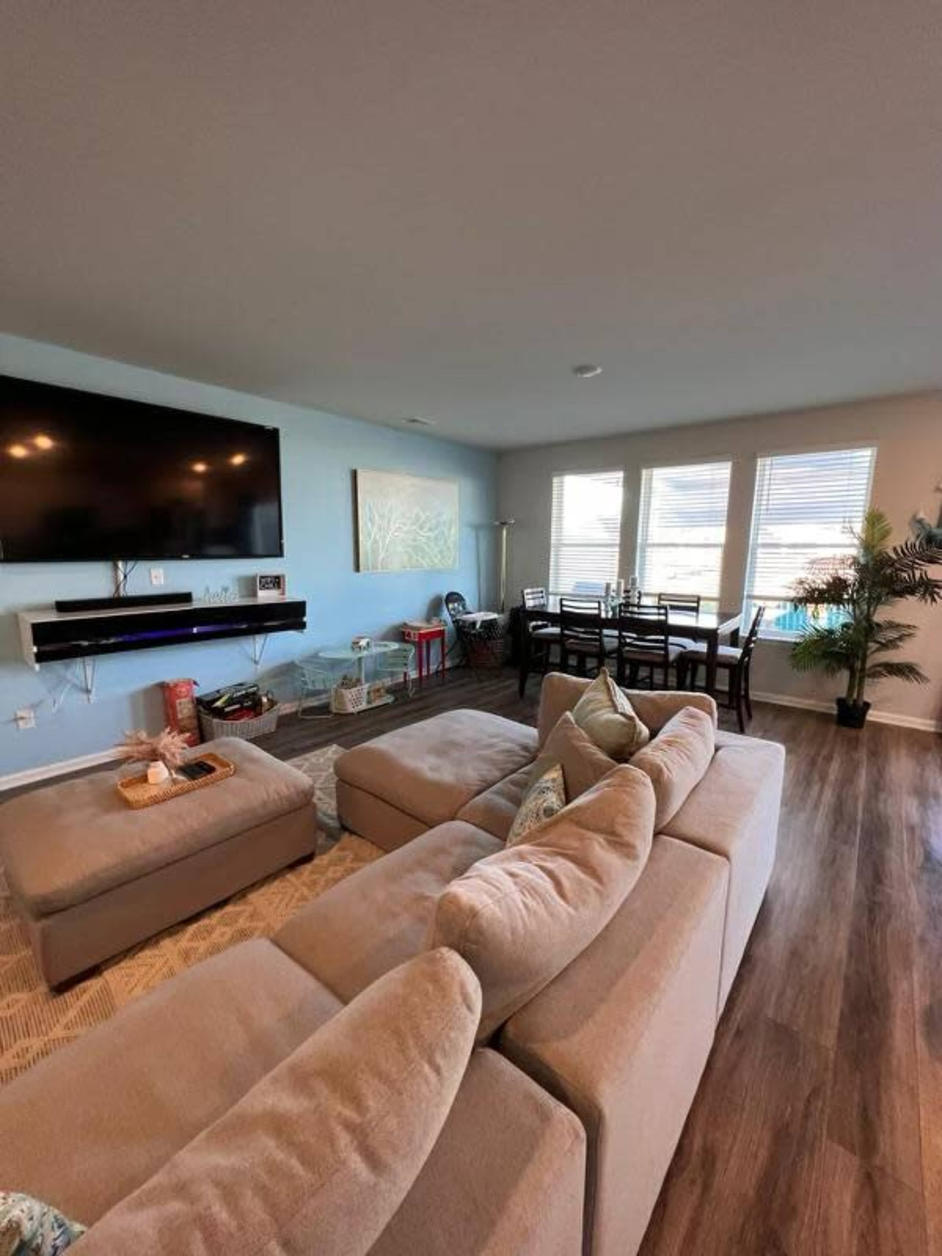 A well-lit living room features a sectional sofa, a large TV mounted on the wall, and a dining area with a table and chairs near the windows.
