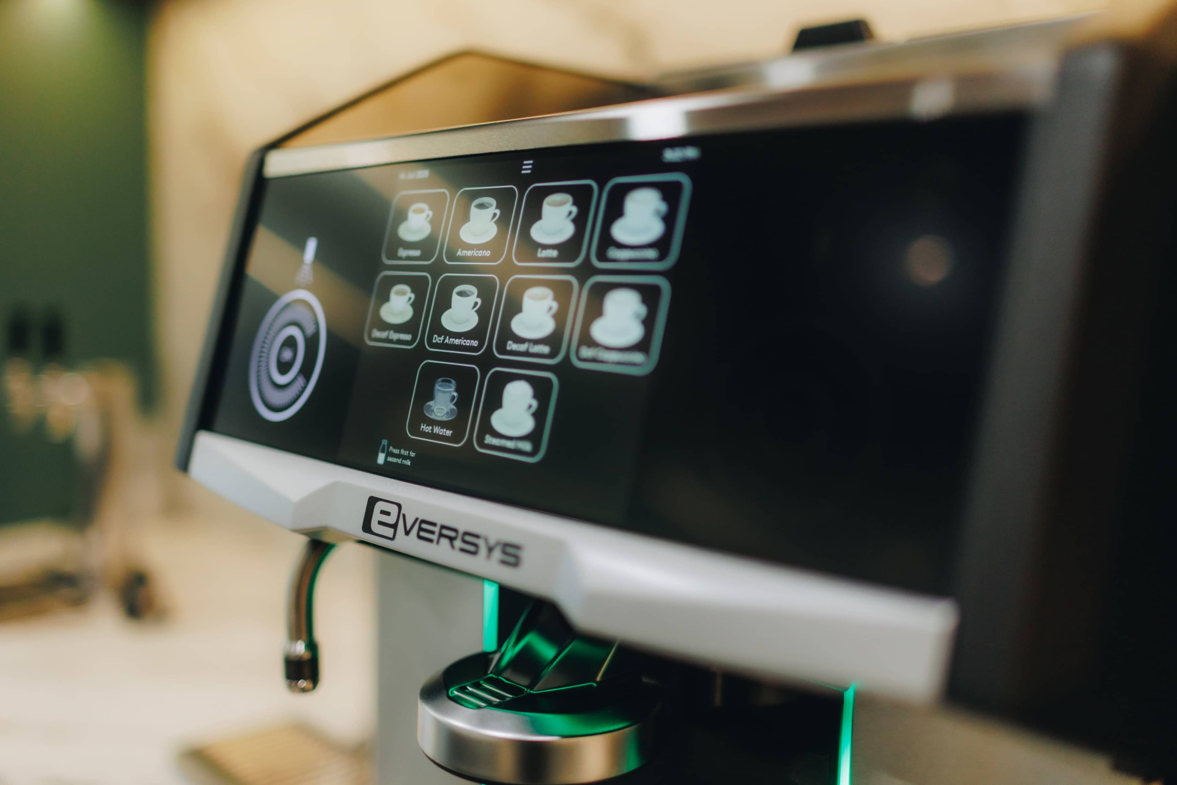 The image shows a close-up of a modern coffee machine with a digital touchscreen interface displaying various beverage options.
