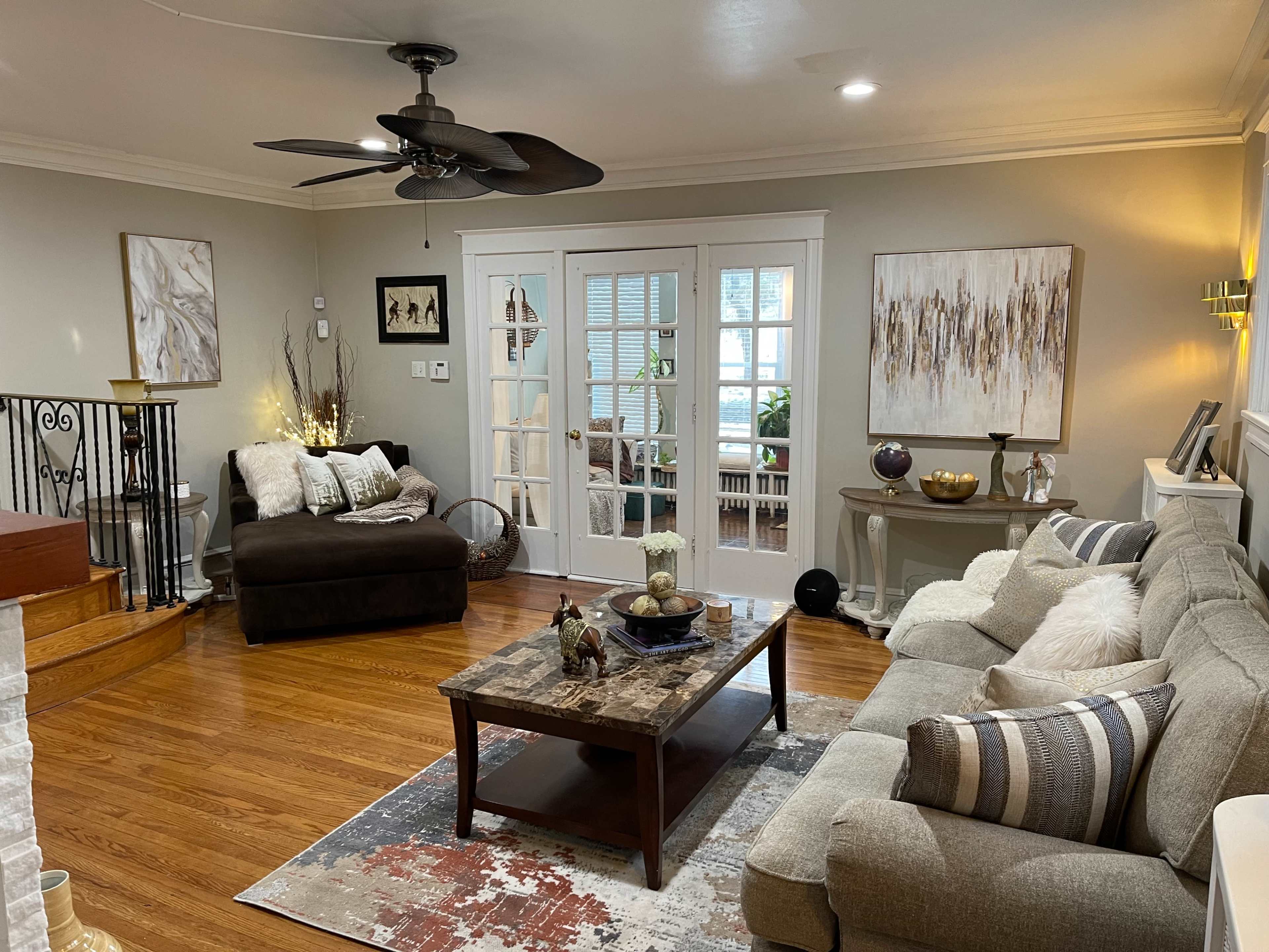 A cozy living room with a sofa, an armchair, a coffee table on a patterned rug, and large windows leading to another room.