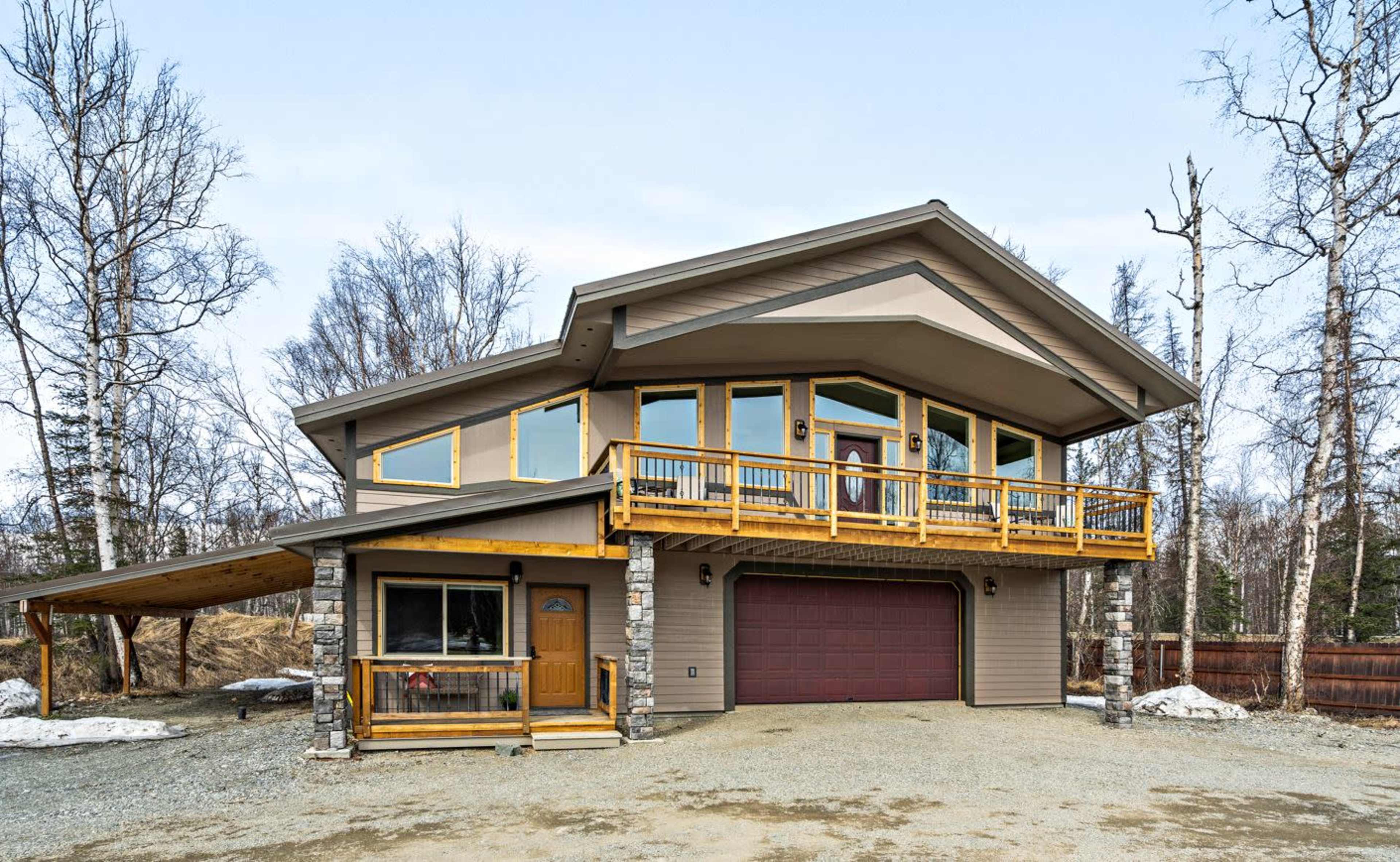 A modern two-story house with large windows, a wooden balcony, and a stone porch, set against a backdrop of bare trees and gravel landscaping.