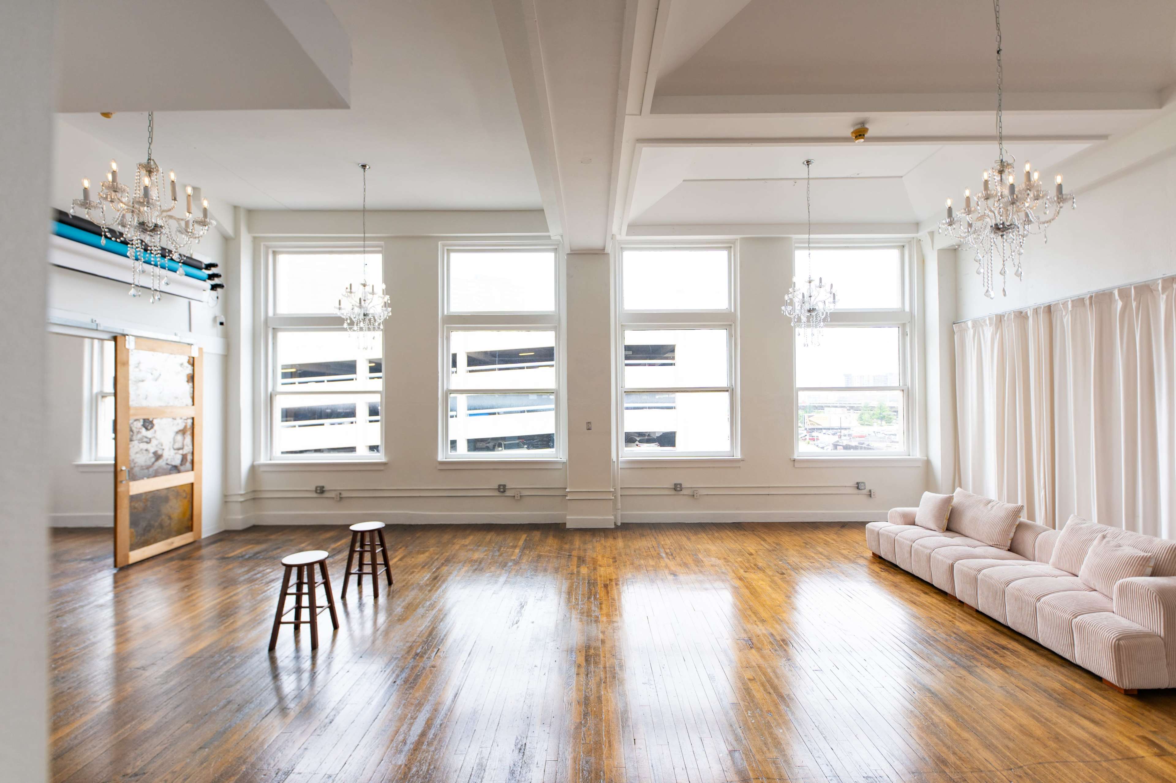 The image shows a spacious, well-lit room with wooden floors, two small stools, and a light-colored sofa along one wall, accented by large windows and chandeliers.