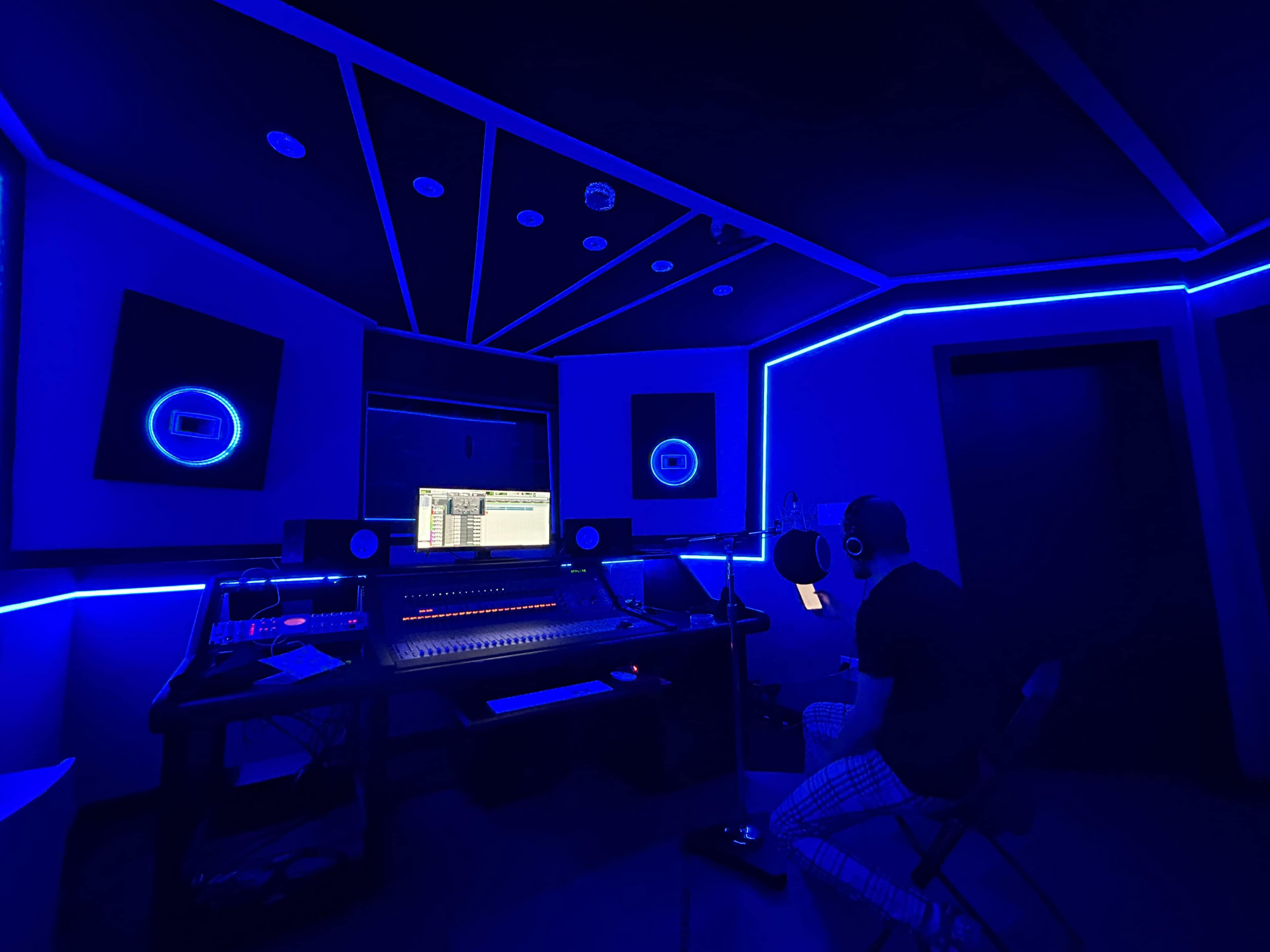 A person sits in a dimly lit recording studio with blue LED lights, facing a computer screen and audio equipment.