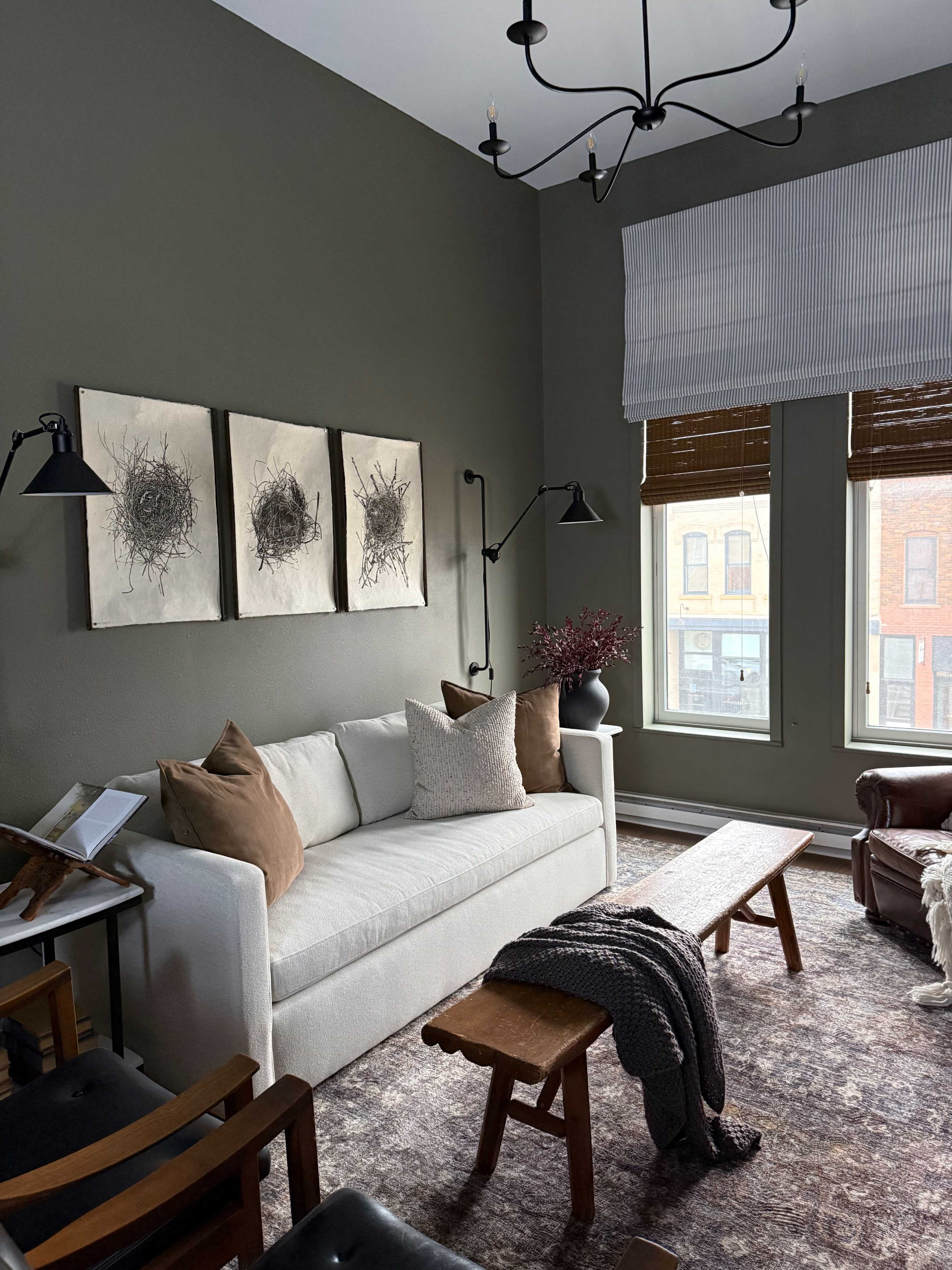 A living room features a white sofa with brown cushions, a wooden bench, and artworks on the green walls, illuminated by natural light from two windows.