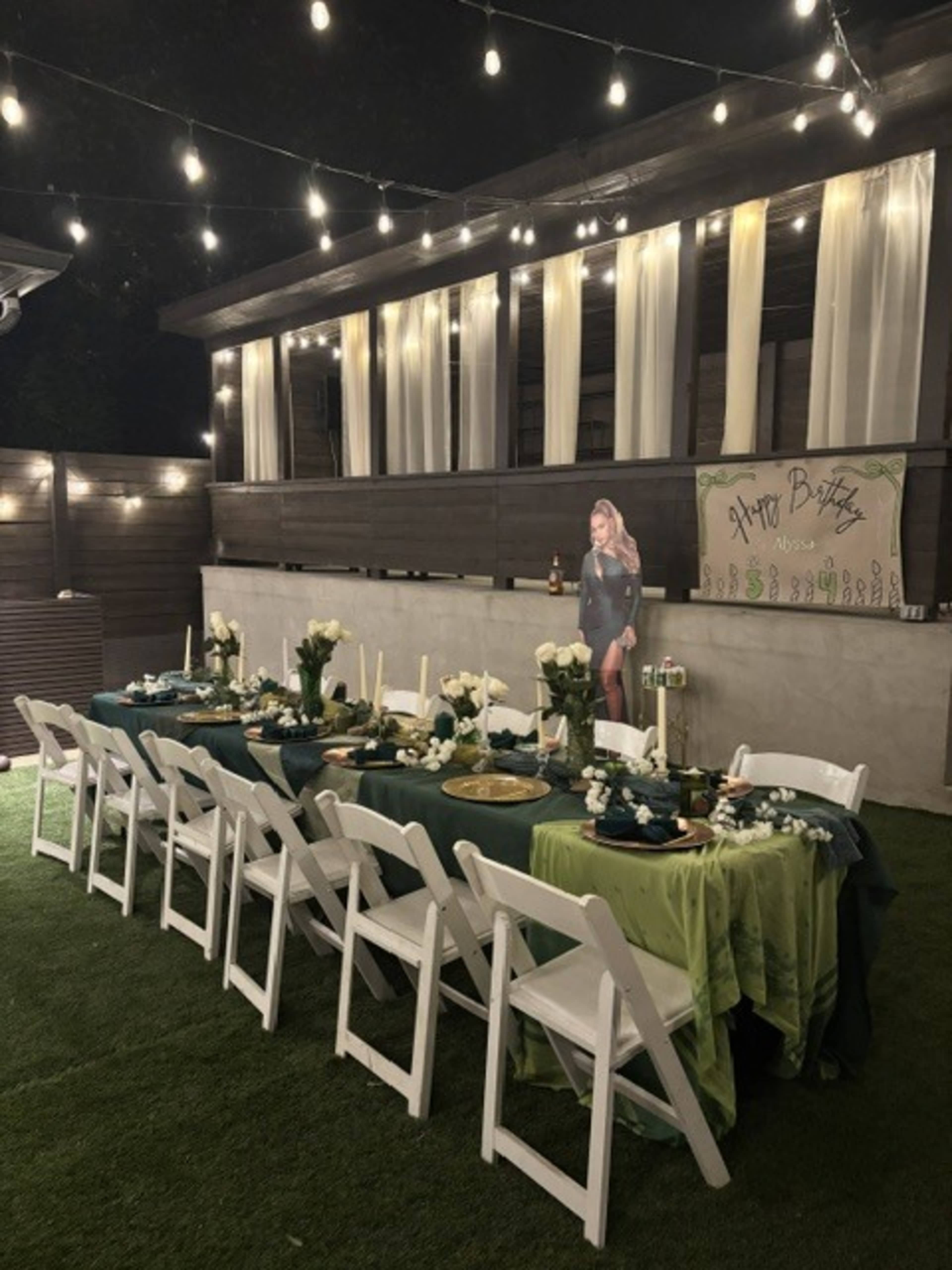 A decorated outdoor venue features a long table set with green and blue tablecloths, white chairs, and floral arrangements, illuminated by string lights.