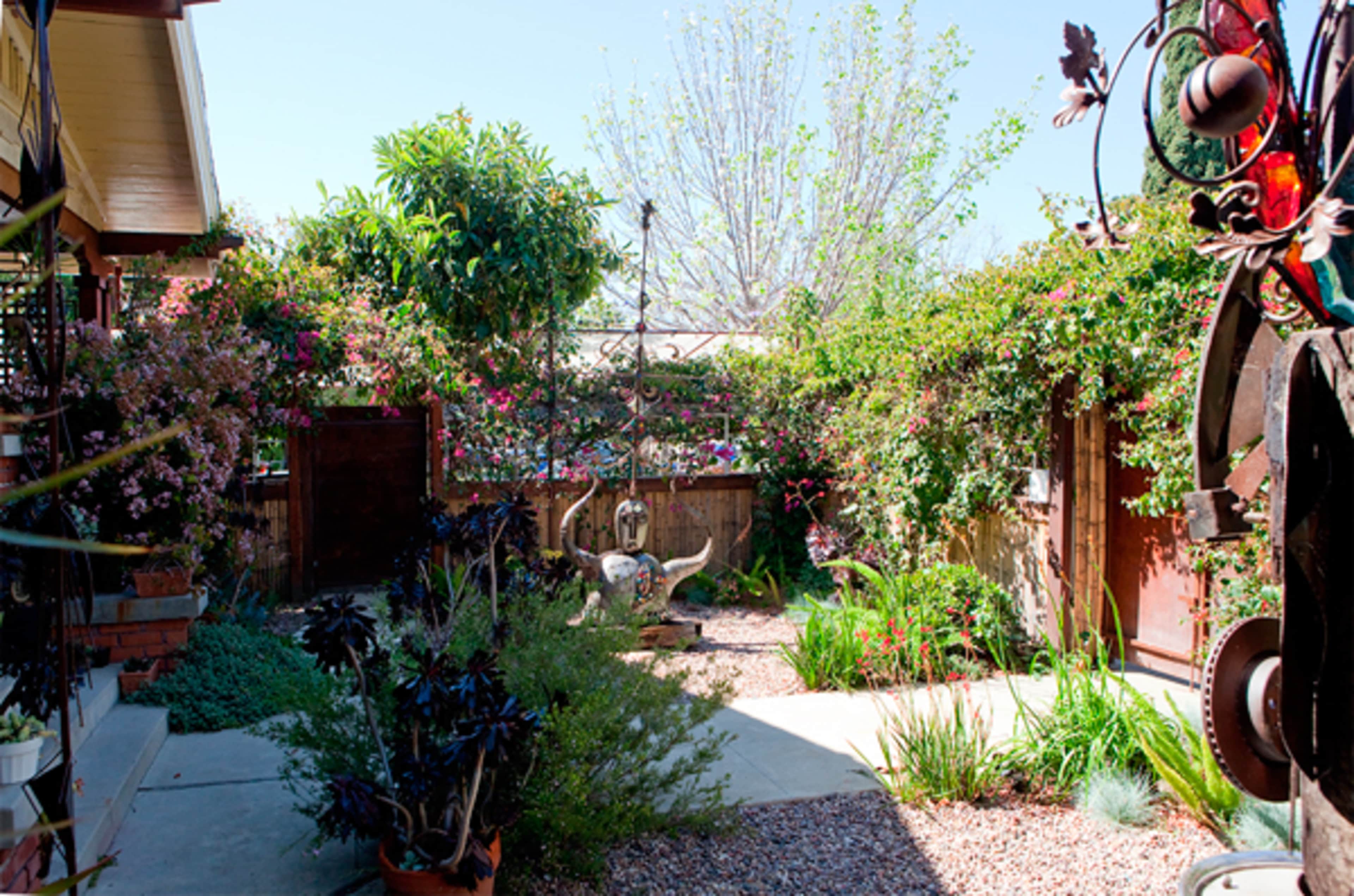 The image shows a landscaped garden area surrounded by flowering plants, decorative sculptures, and a pathway leading through the space.