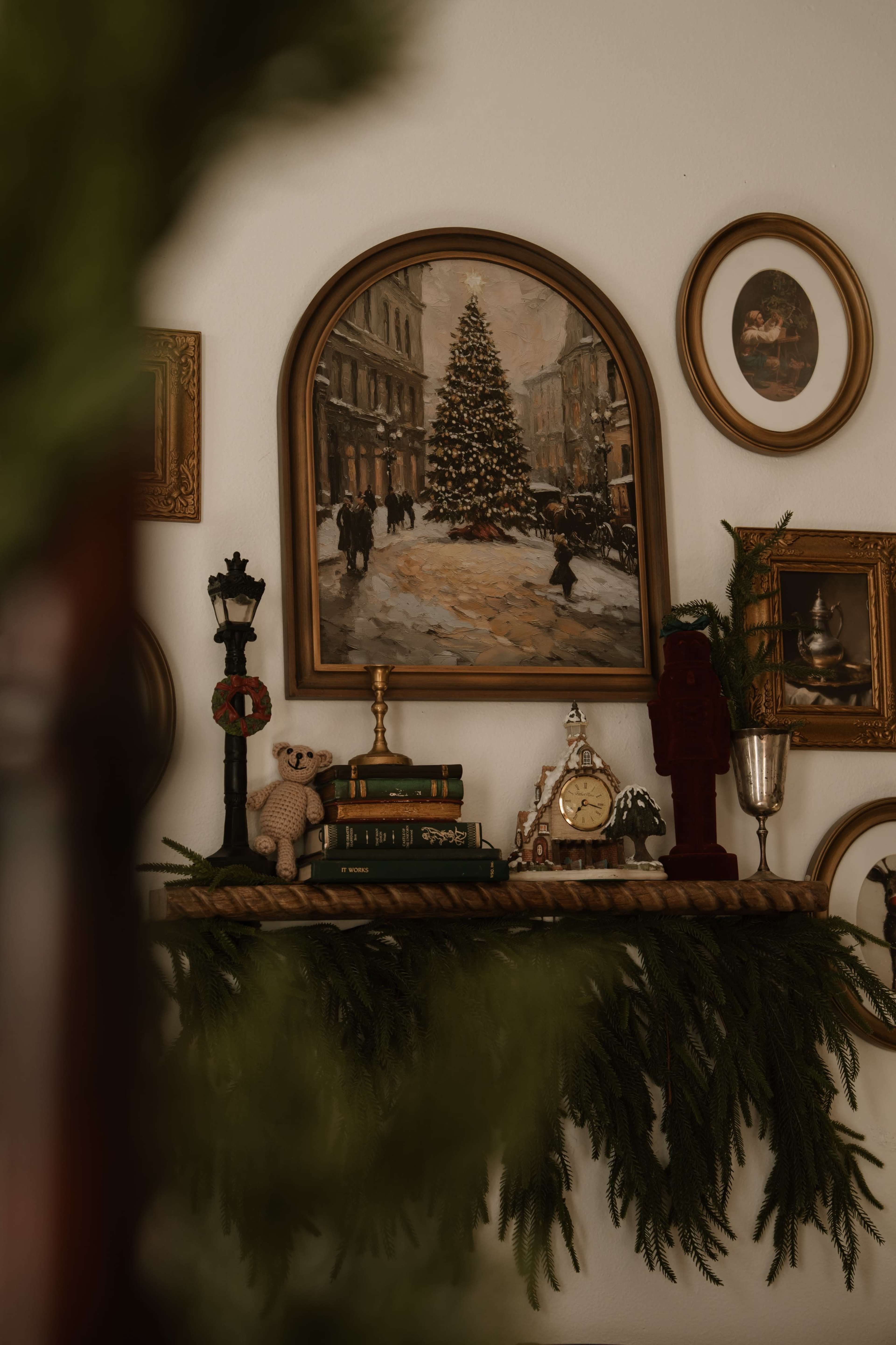 The image features a shelf adorned with a vintage painting of a Christmas tree, surrounded by decorative frames and various festive items, including a clock and a teddy bear.
