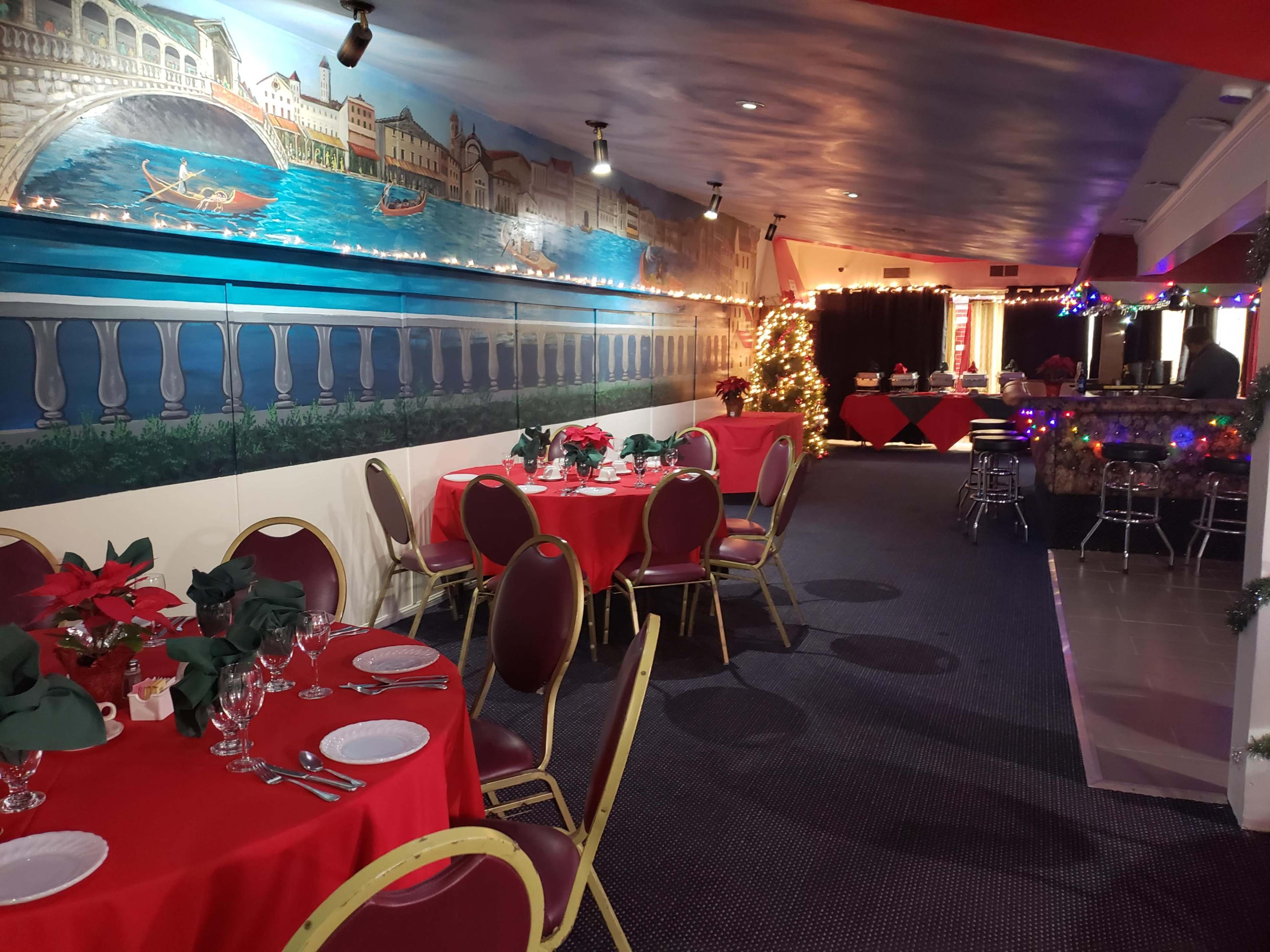 The image showcases a decorated banquet hall featuring round tables with red tablecloths, adorned with festive centerpieces, and a mural depicting a waterfront scene.