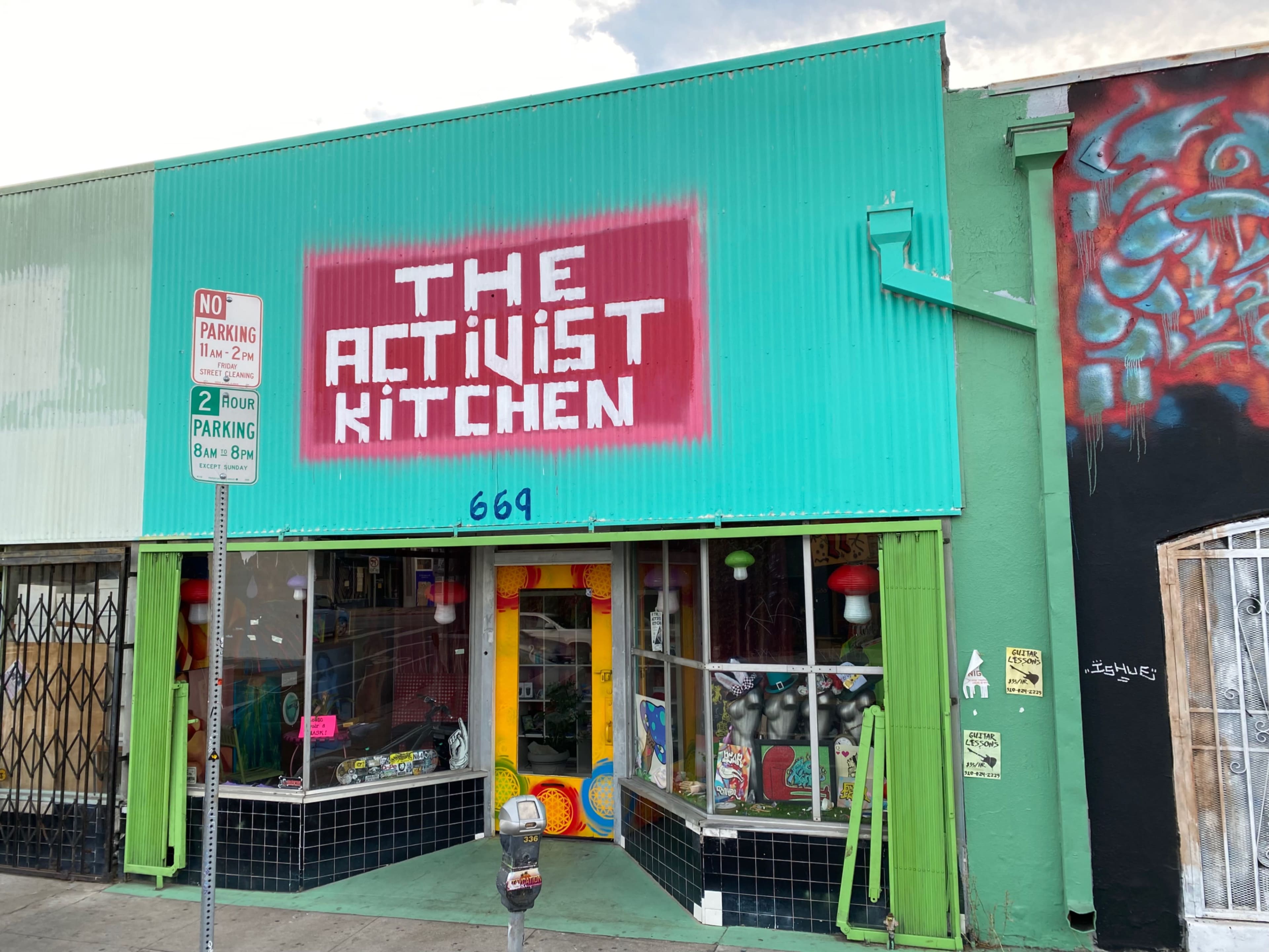 A brightly colored storefront with the sign "THE ACTIVIST KITCHEN" prominently displayed above a window featuring colorful decorations.