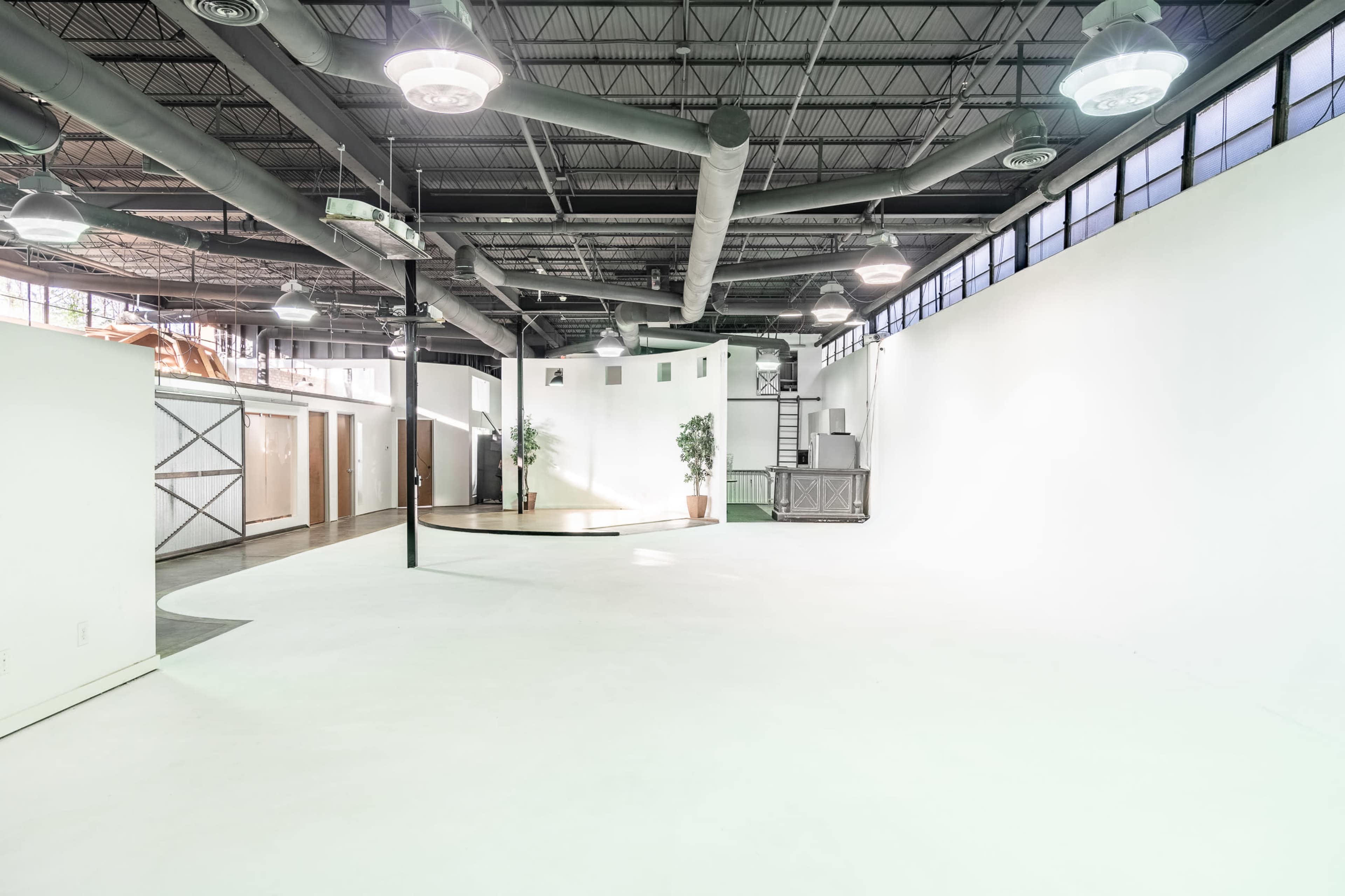 The image shows a spacious, empty interior with white walls, a polished floor, and large windows, featuring minimalist design elements and some greenery.