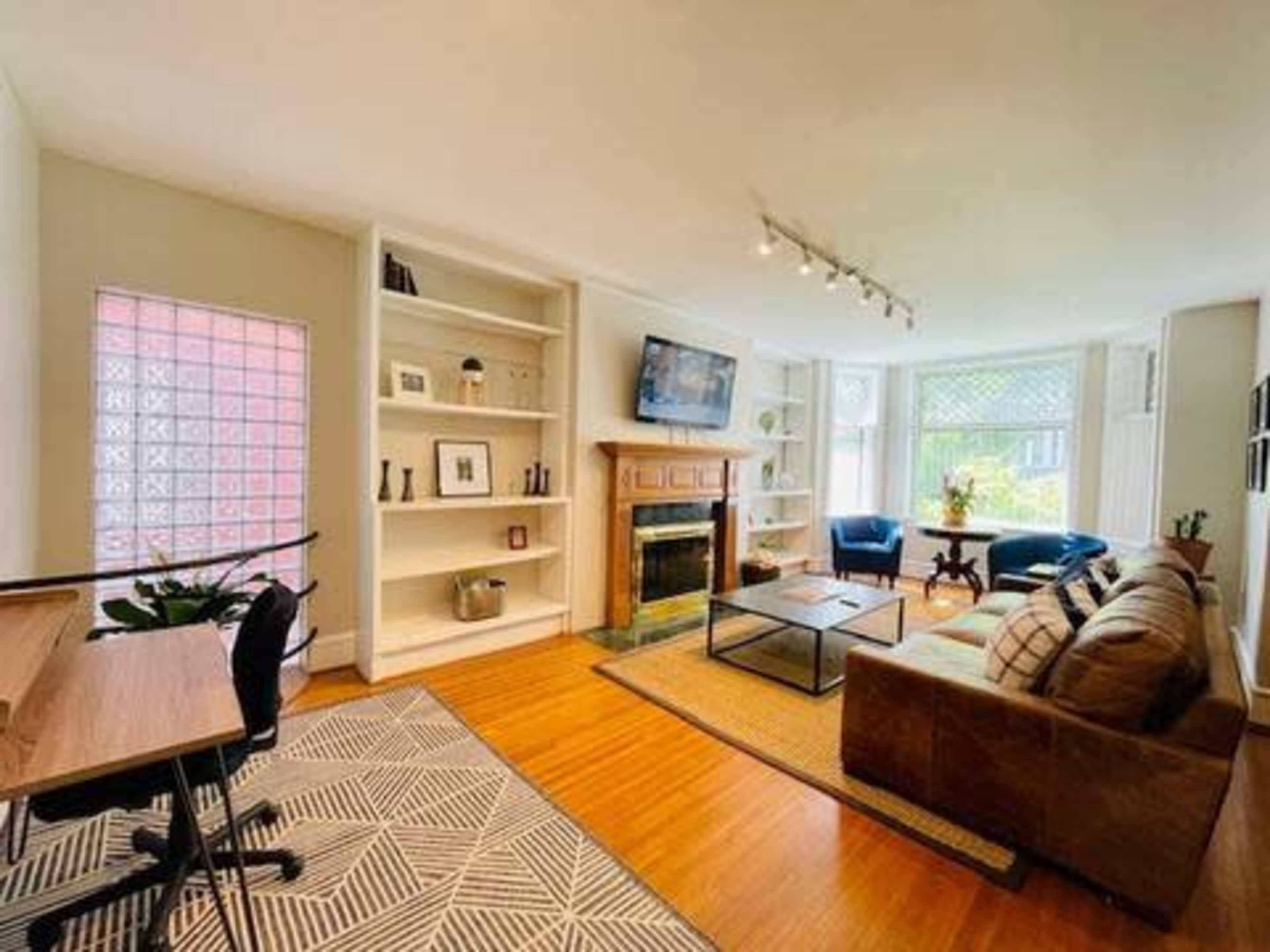 The image shows a well-lit living room with a fireplace, a television mounted on the wall, a cozy seating area, a desk, and a large window for natural light.