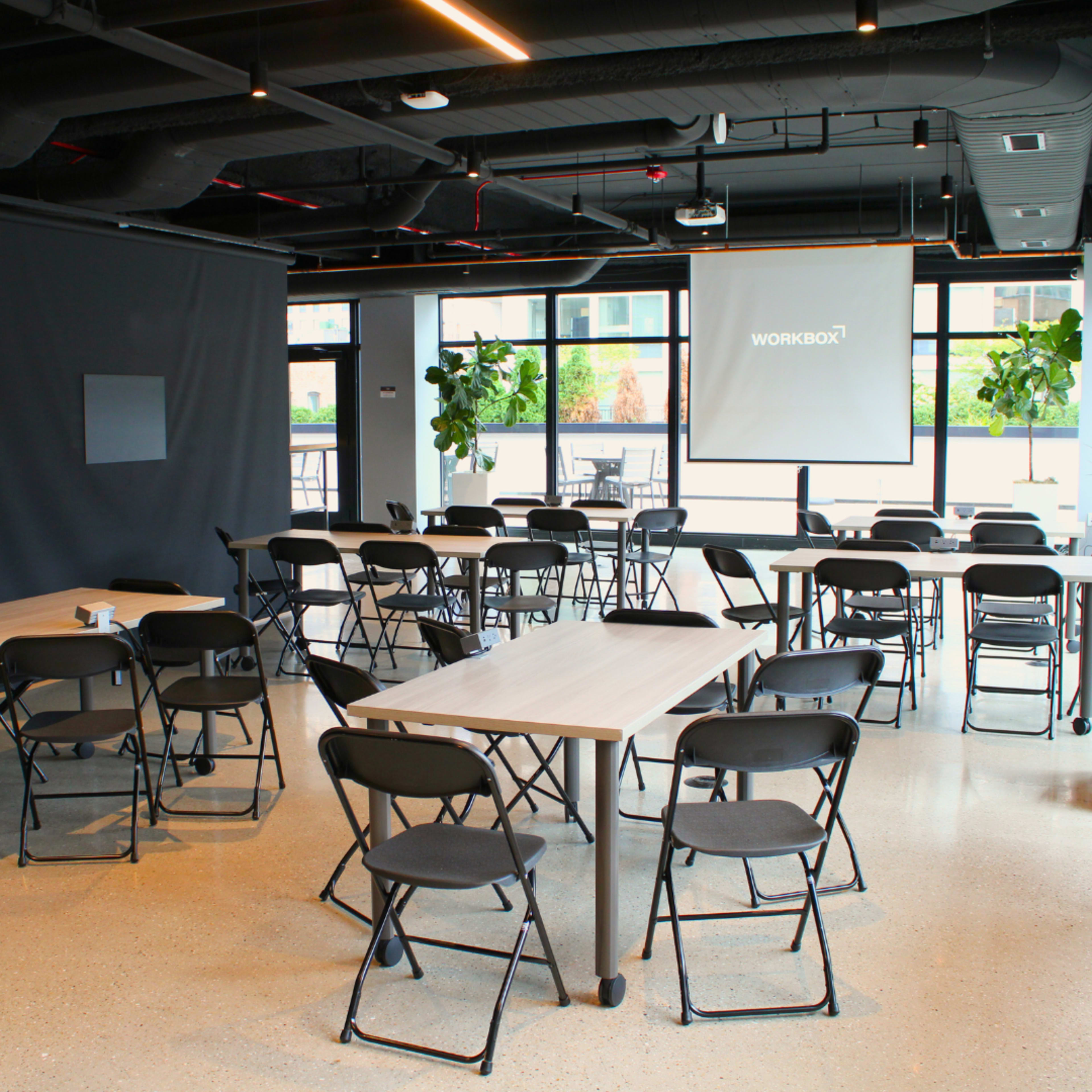 The image shows a modern conference room with several tables and black folding chairs arranged for a meeting, along with a projector screen displaying the word "WORKBOX."