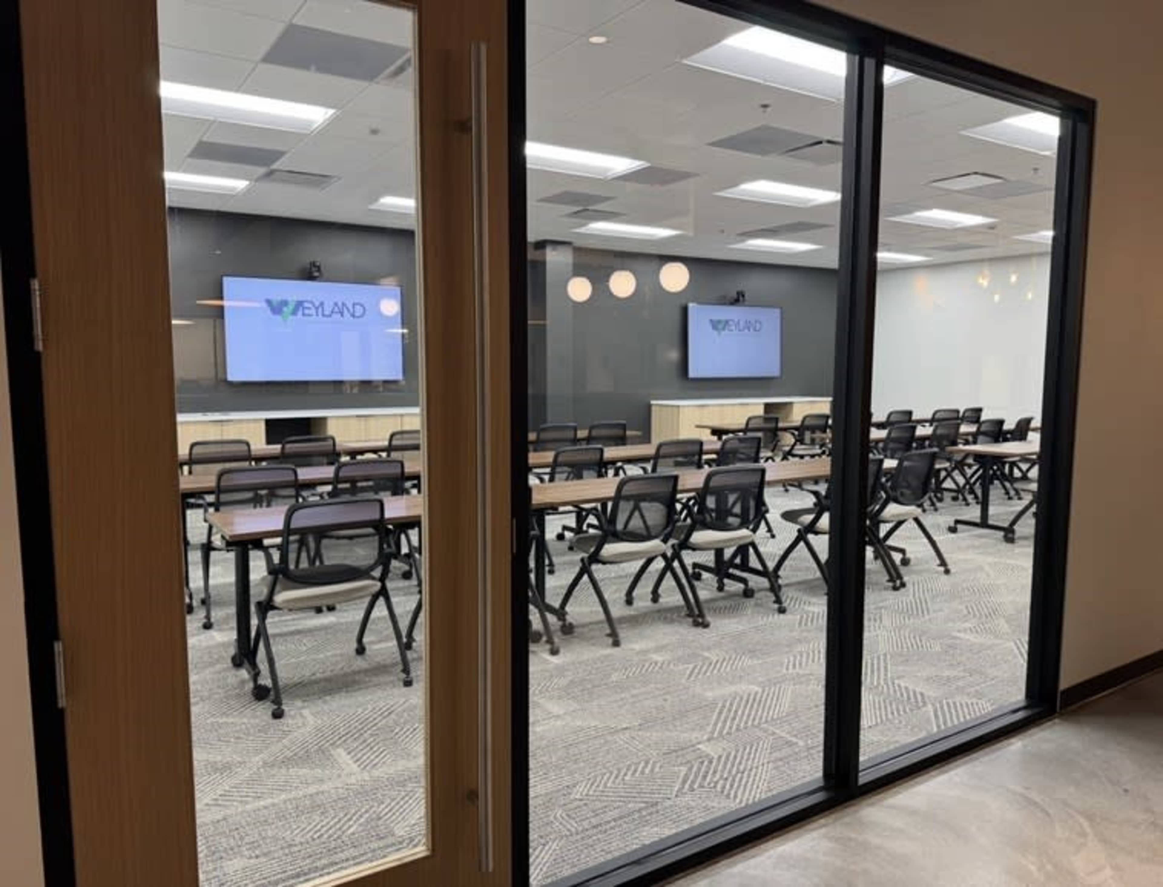 The image shows a conference room with rows of chairs and tables, equipped with two monitors displaying the name "Wayland."