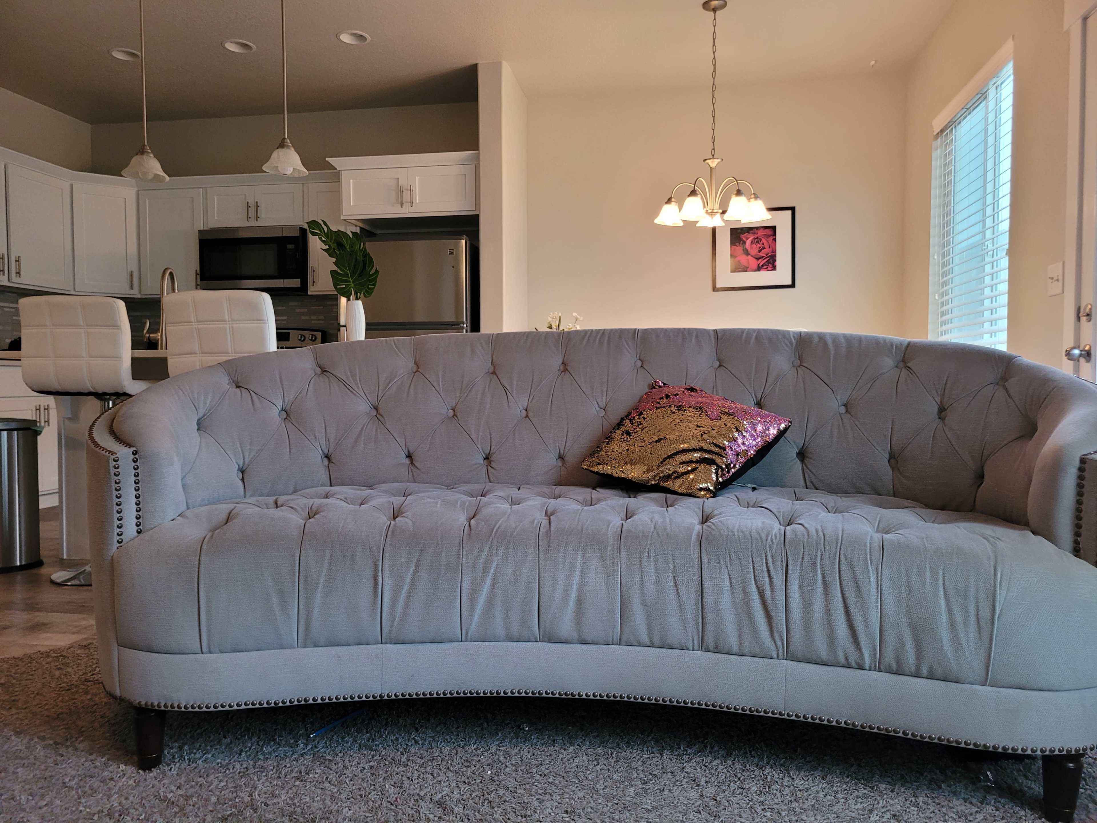 A gray tufted couch with a decorative pillow sits in a well-lit living area, facing a kitchen with white cabinetry and modern fixtures.