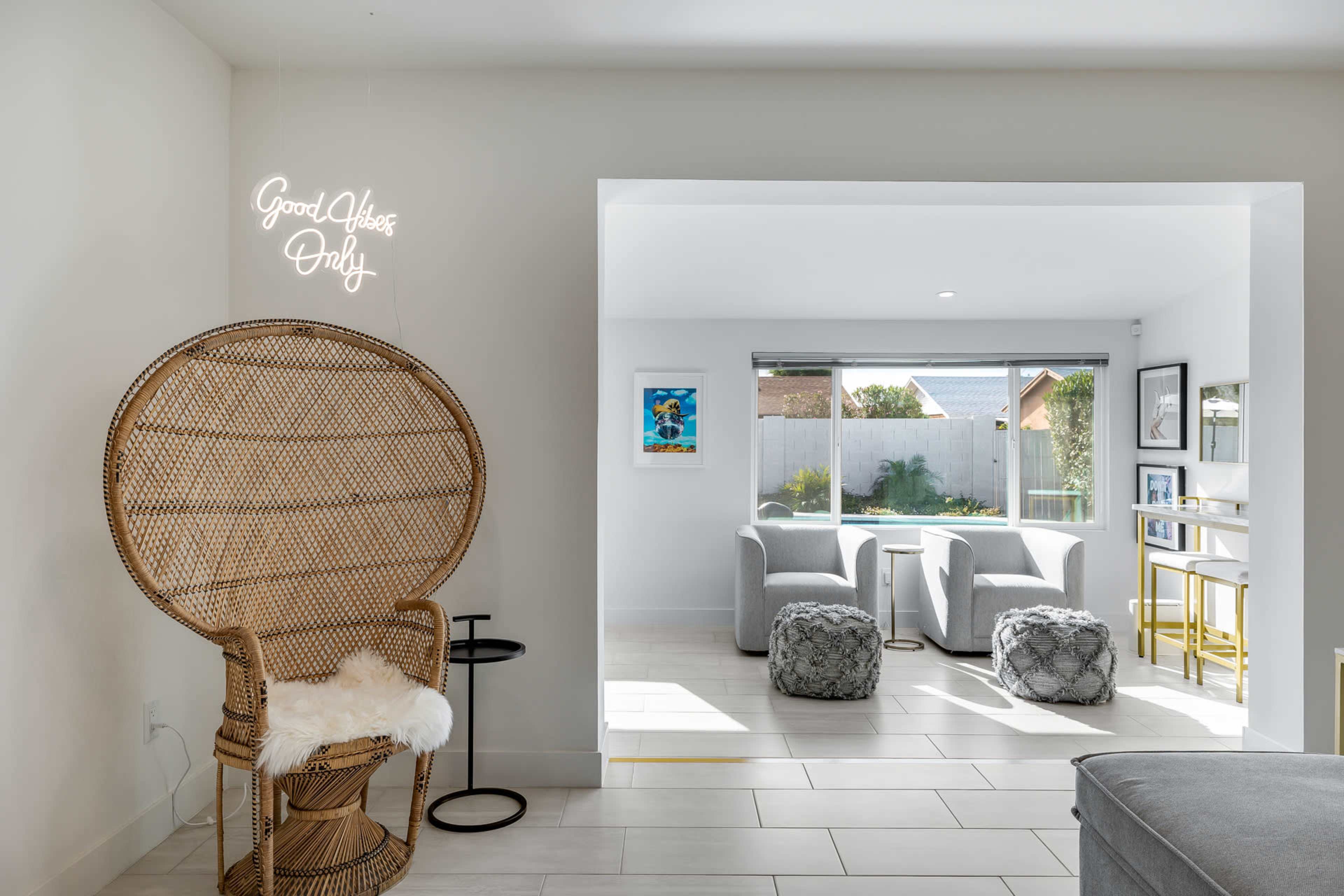 A cozy living area features a rattan chair with a "Good Vibes Only" sign, two armchairs, and a small table in a well-lit space with modern decor.