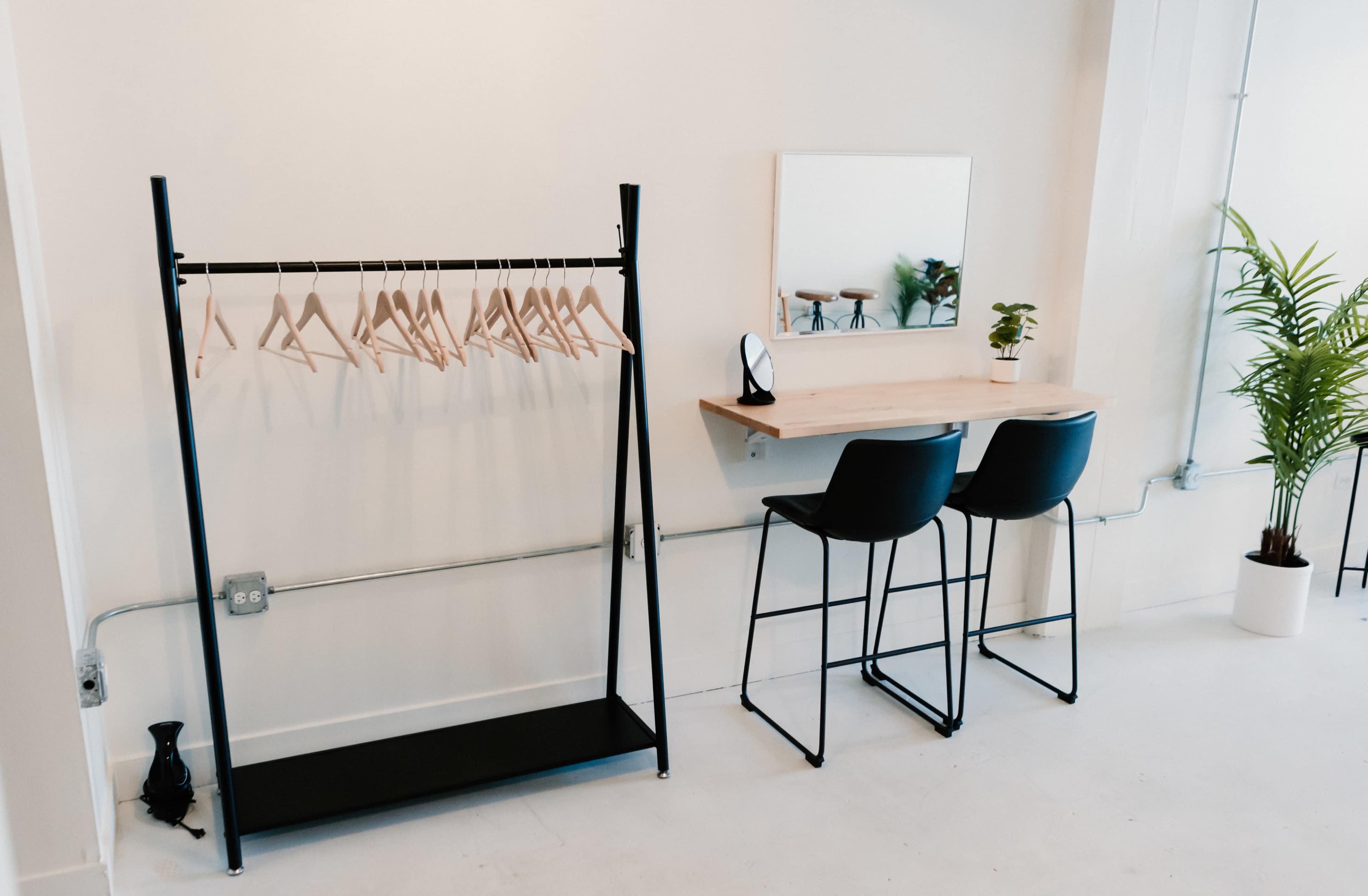A minimalist interior featuring a clothing rack with wooden hangers, a small table with two black chairs, and a potted plant.
