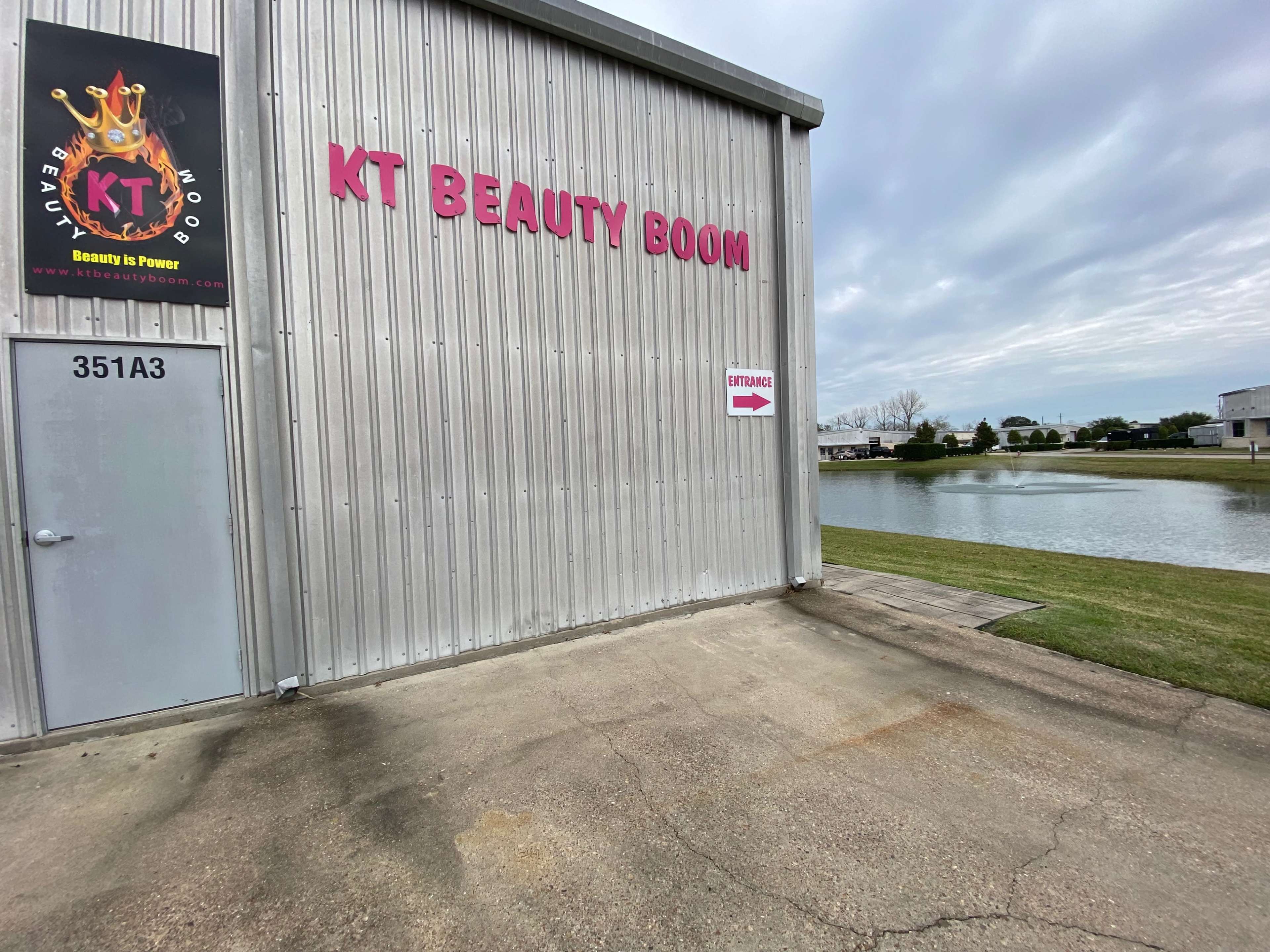 A metal building with a sign reading "KT BEAUTY BOOM" and an arrow pointing toward the entrance, situated next to a calm body of water.