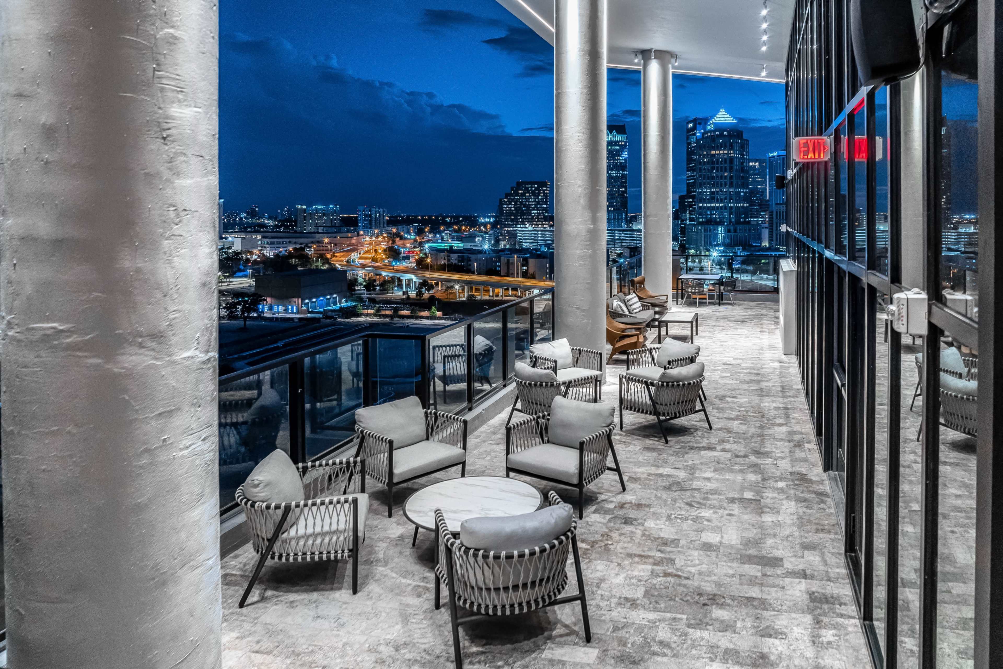 A modern balcony features several chairs and tables overlooking a city skyline at dusk.