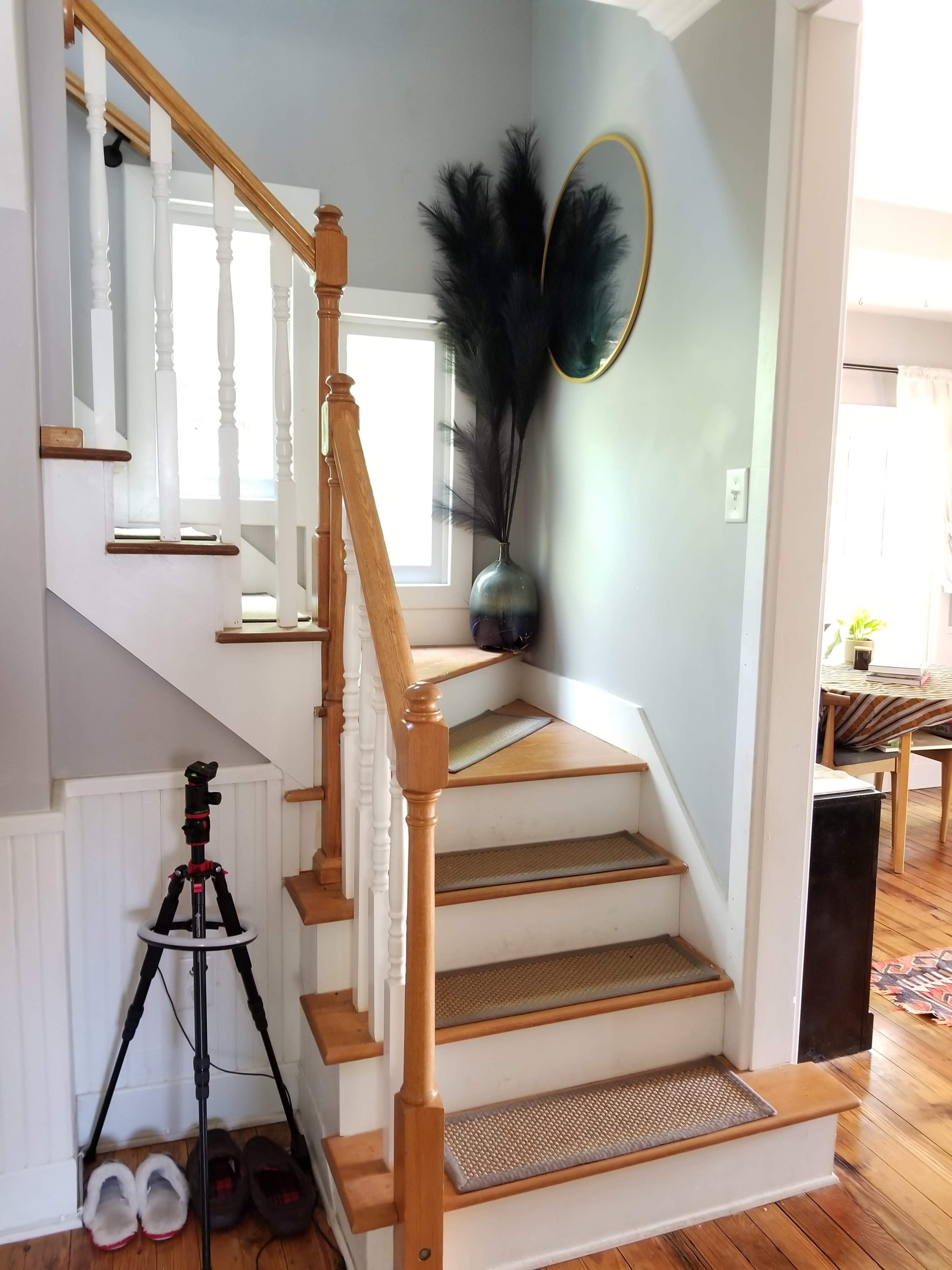 A wooden staircase leads to an upper level, flanked by a round mirror and a decorative vase with tall, dark foliage.