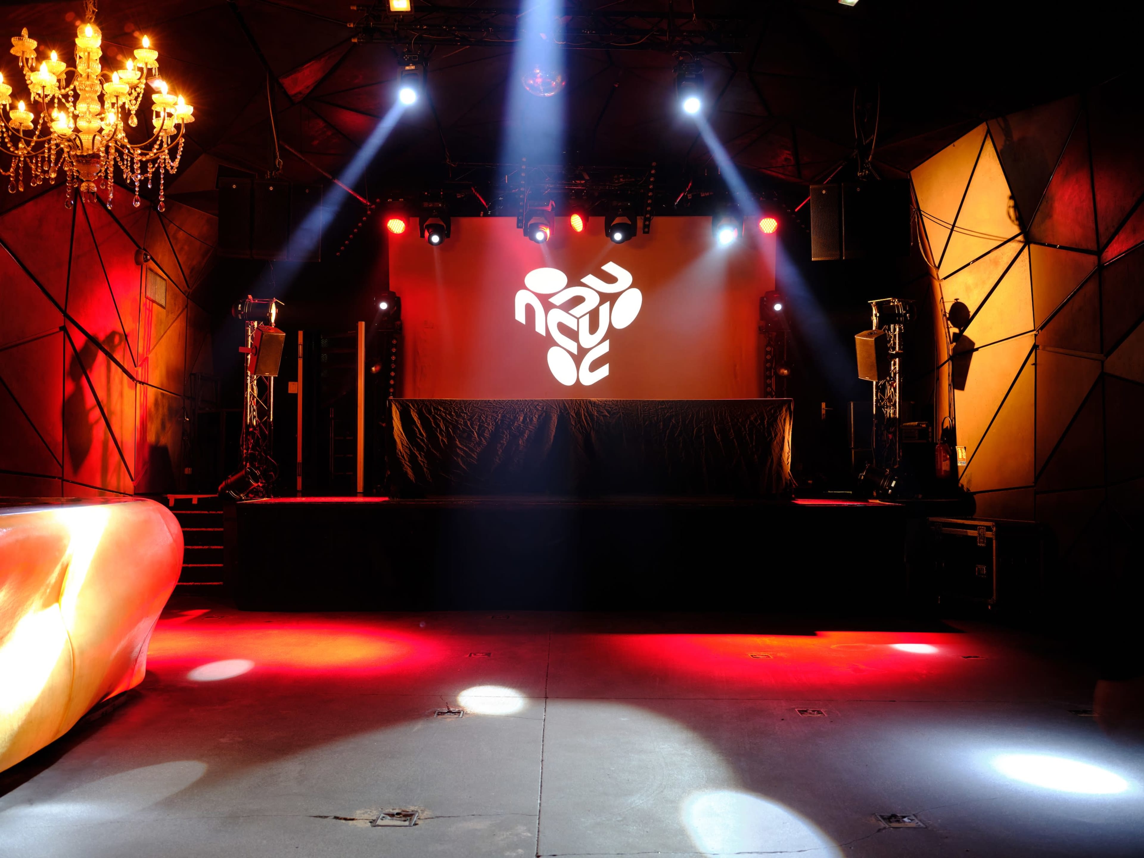 The image shows an illuminated stage inside a venue with a DJ setup, colorful lighting, and geometric wall designs.