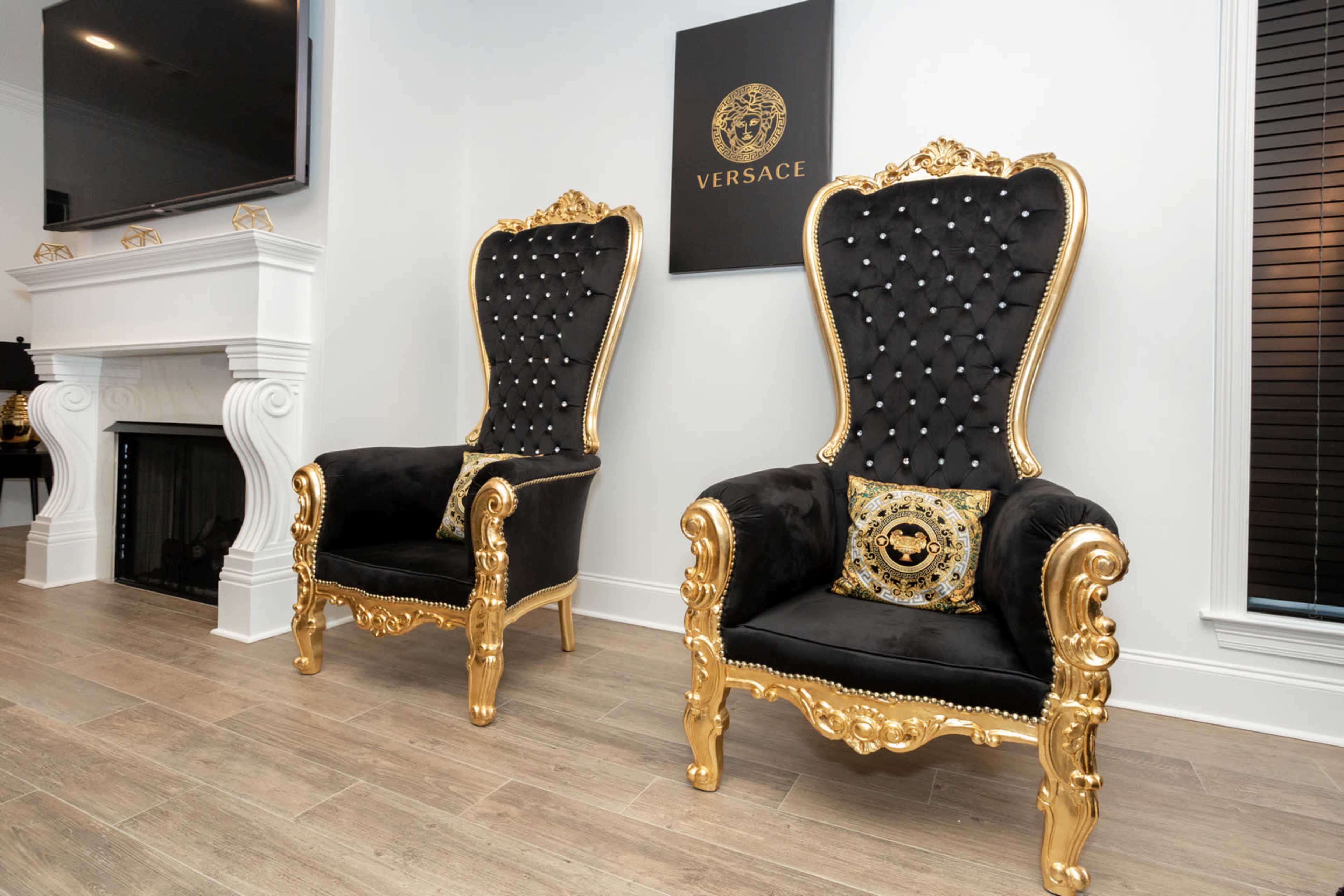 Two ornate black and gold armchairs with tufted upholstery placed next to a fireplace and a wall adorned with a Versace logo.
