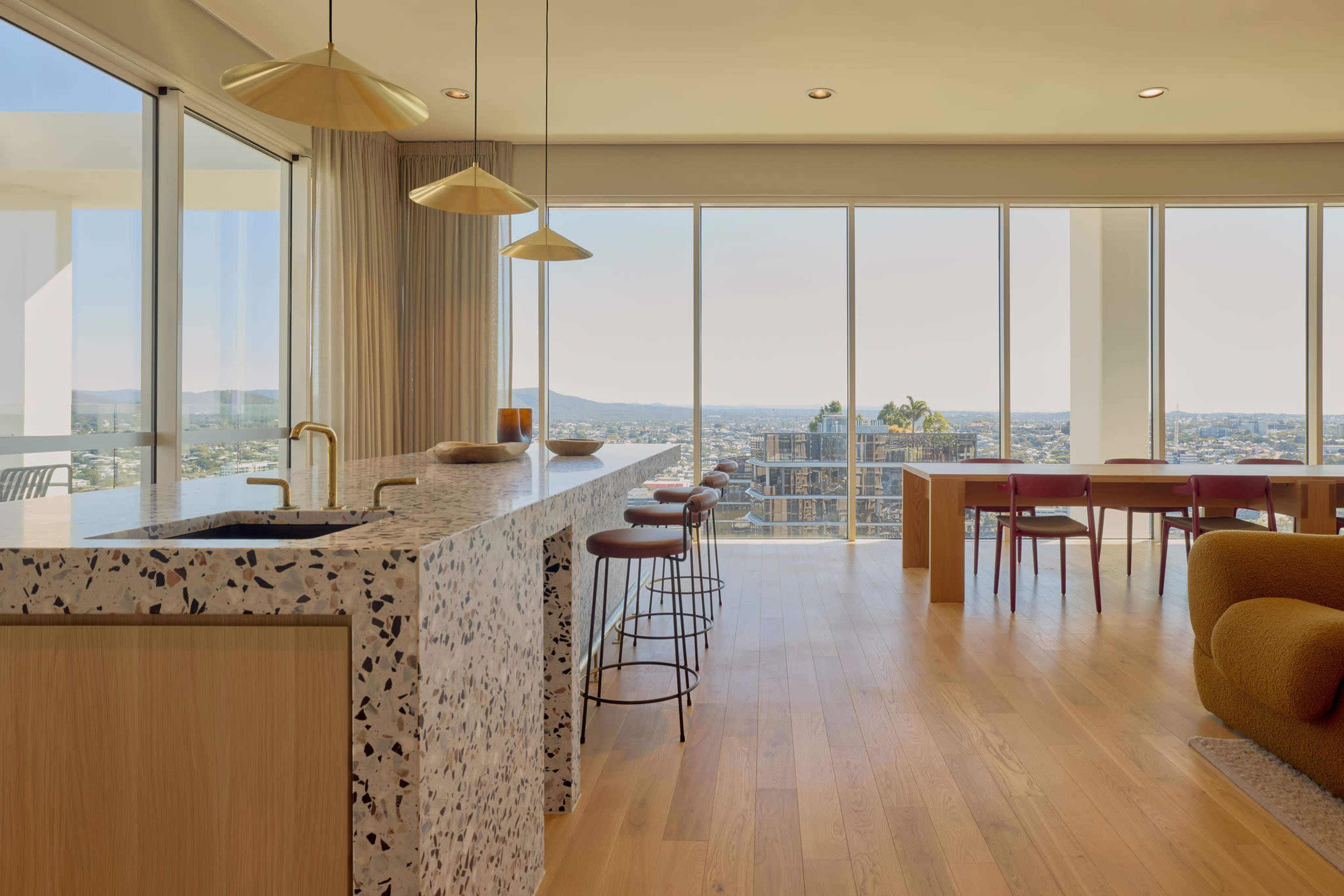 A modern kitchen and dining area are illuminated by natural light, featuring a terrazzo countertop and expansive city views through large glass windows.