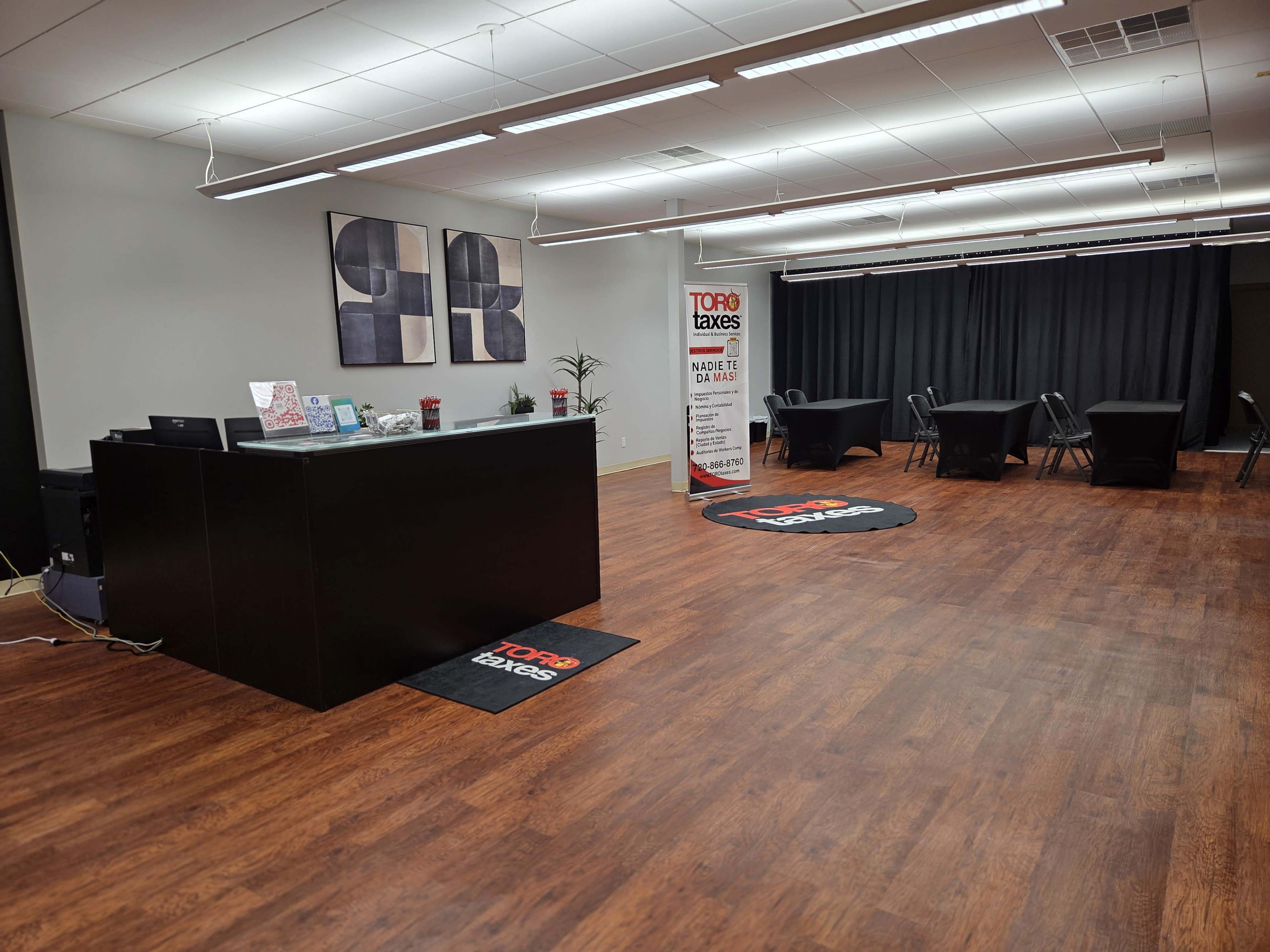 The image shows a spacious office interior with a reception area featuring a black counter, promotional materials, and a seating space with black tables and chairs.