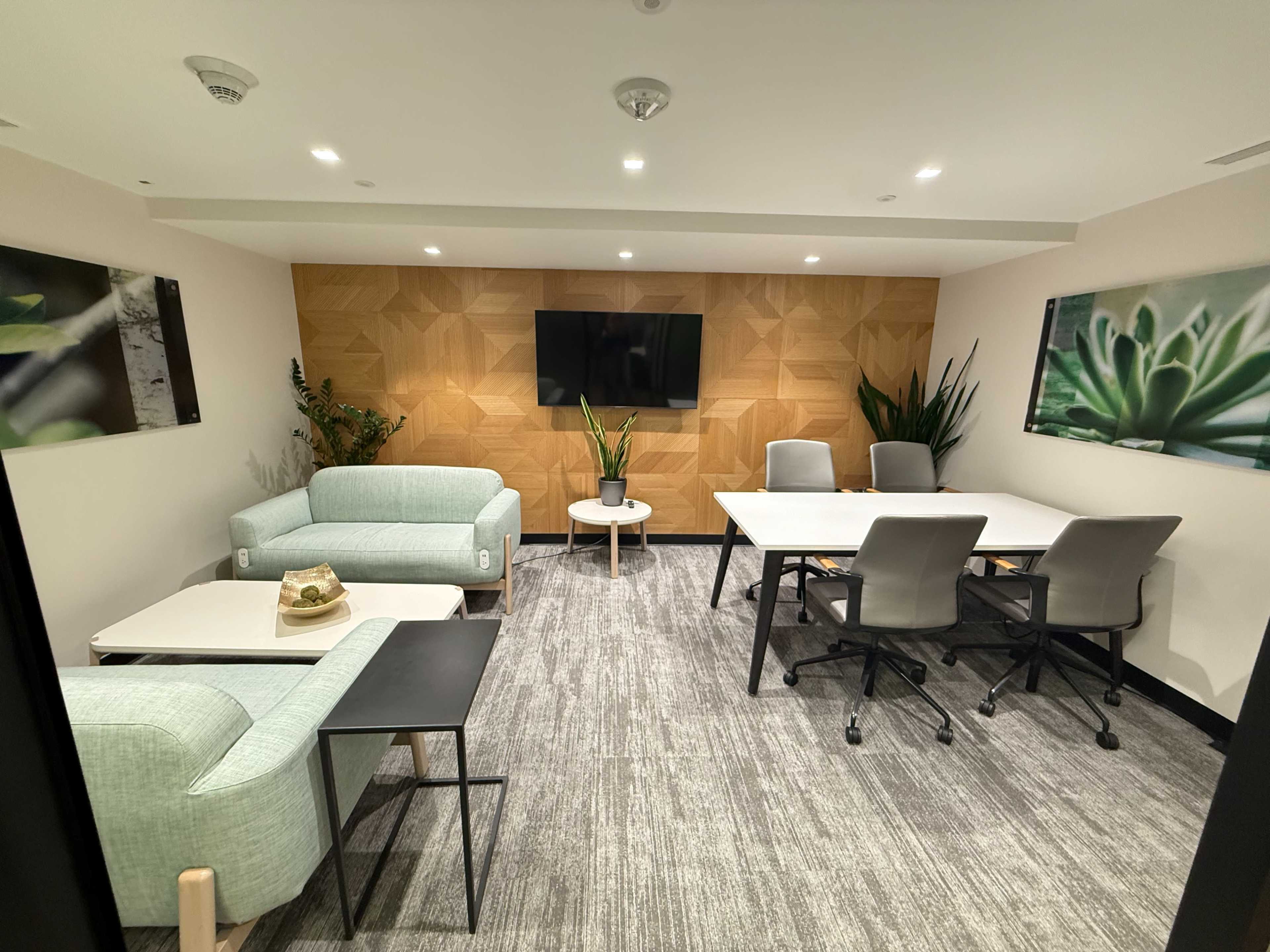 The image shows a modern meeting room with light green sofas, a large table surrounded by chairs, and two plant-themed artworks on the walls.
