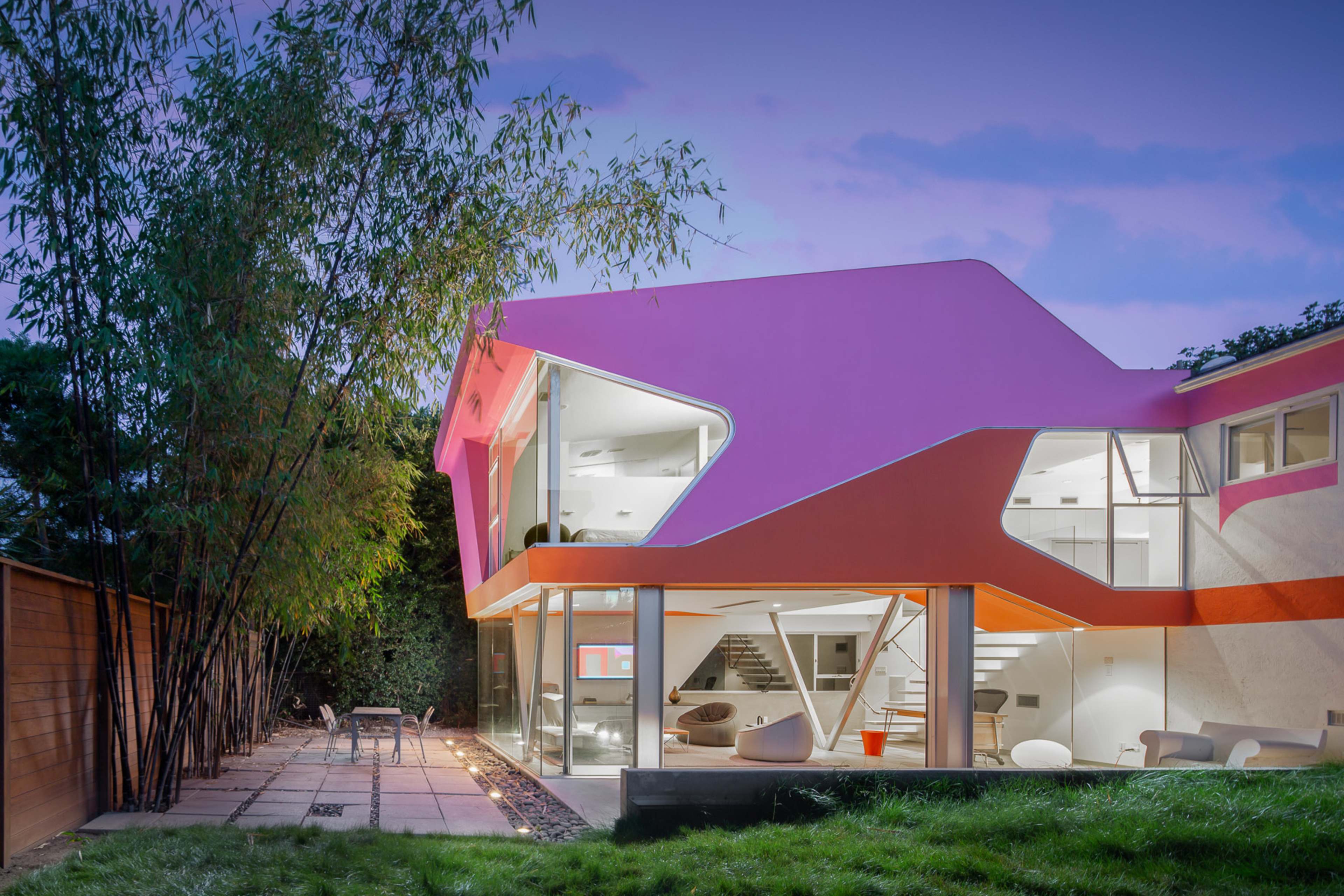 A modern house with a distinctive pink and orange exterior, featuring large glass windows and a unique angular design, surrounded by greenery and a patio area.