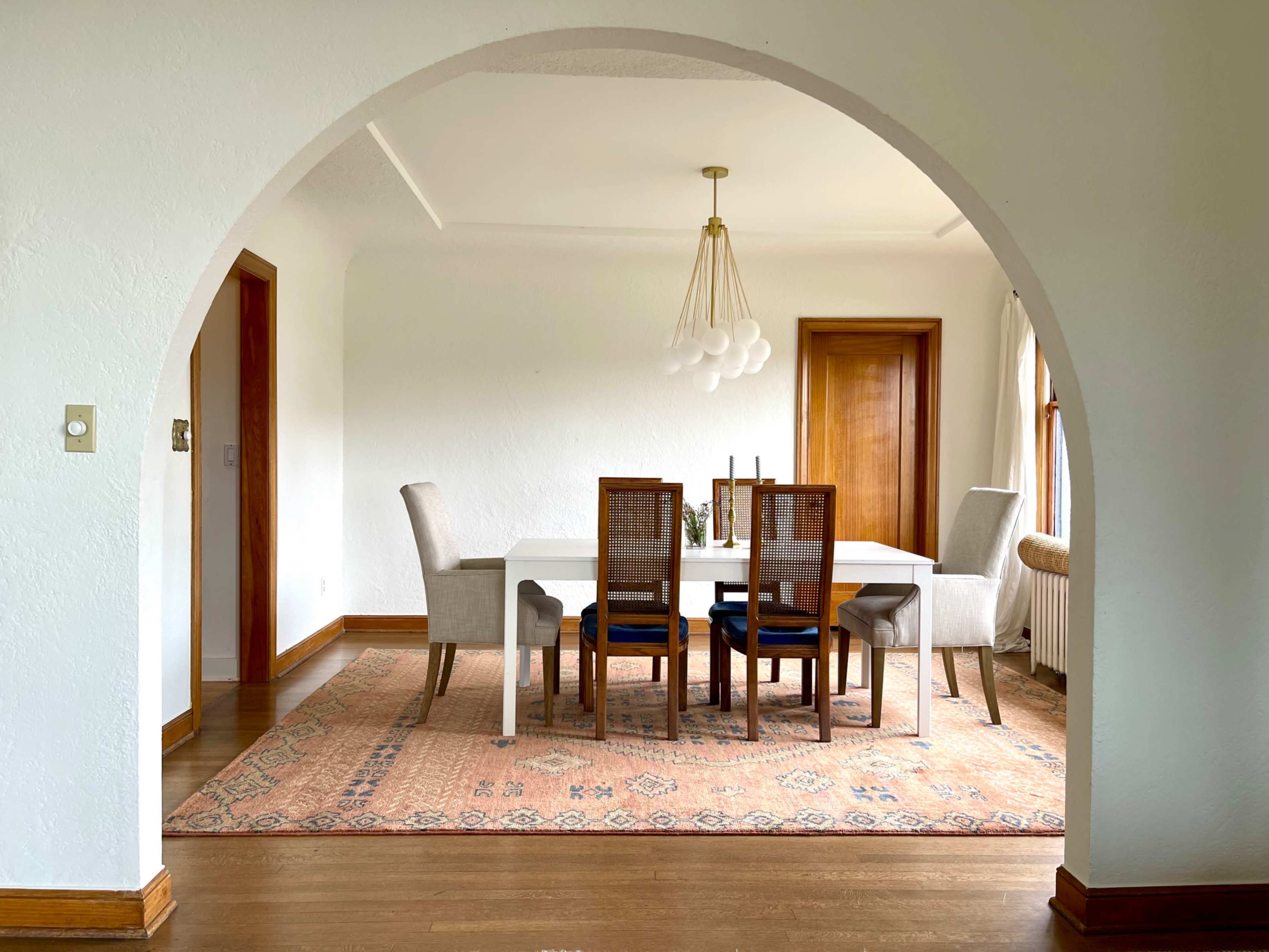A dining room features a white table surrounded by six chairs, with a decorative rug beneath and an arched entrance.
