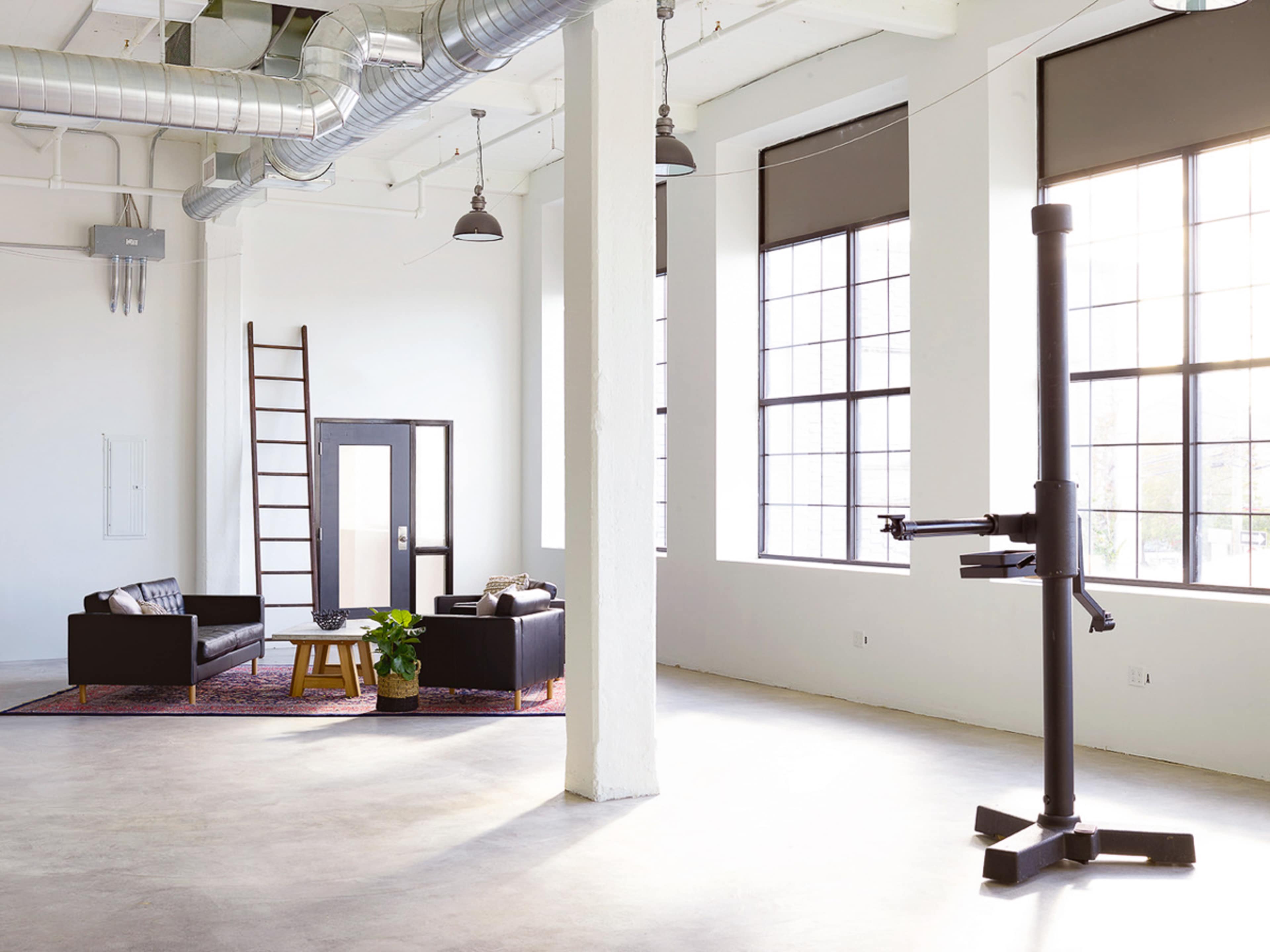 The image shows a spacious, modern interior with large windows, a ladder against the wall, and a seating area featuring black furniture and a small plant.