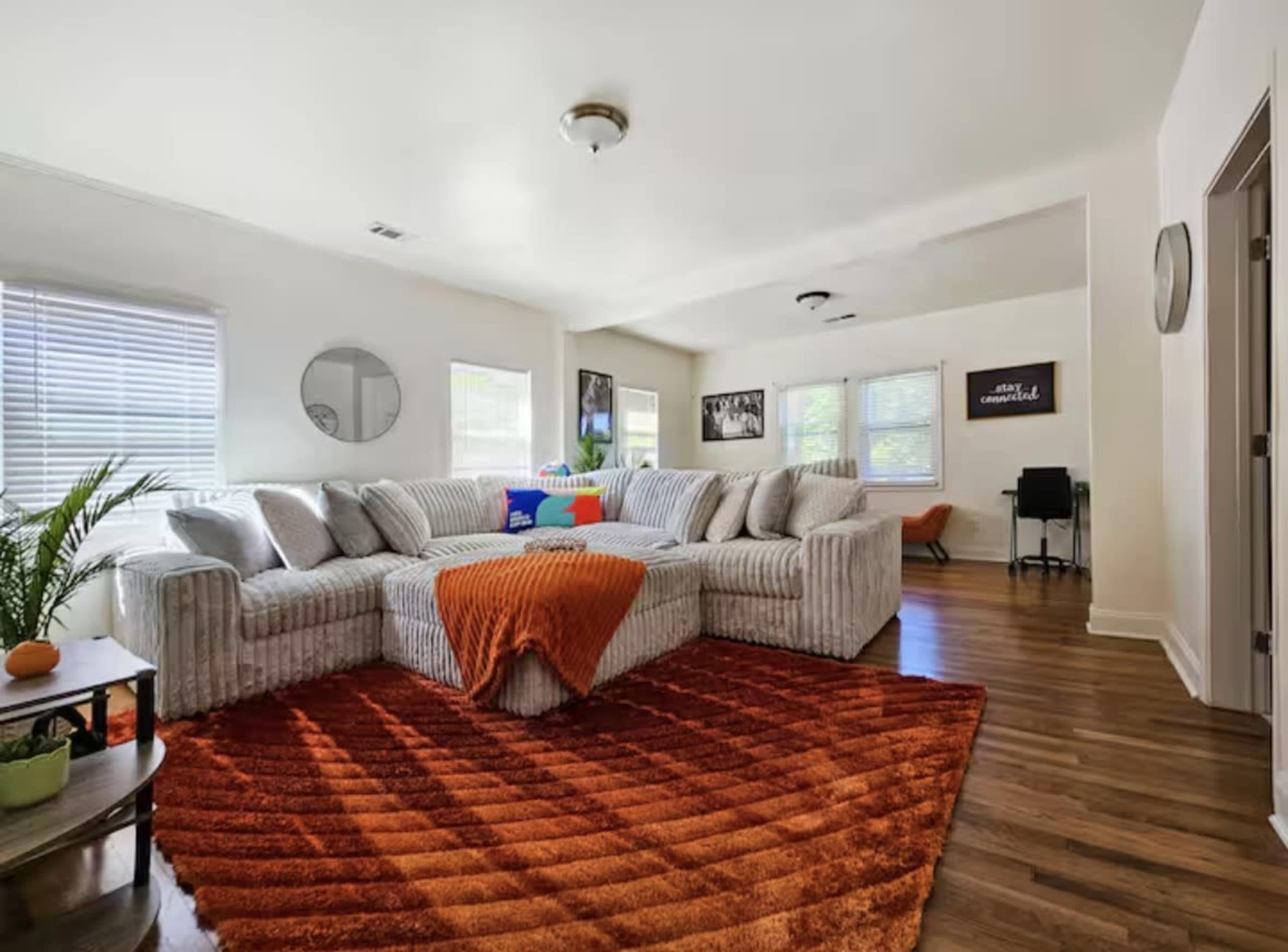 The image shows a living room with a large sectional couch, an orange rug, wooden floors, and various decorative items.