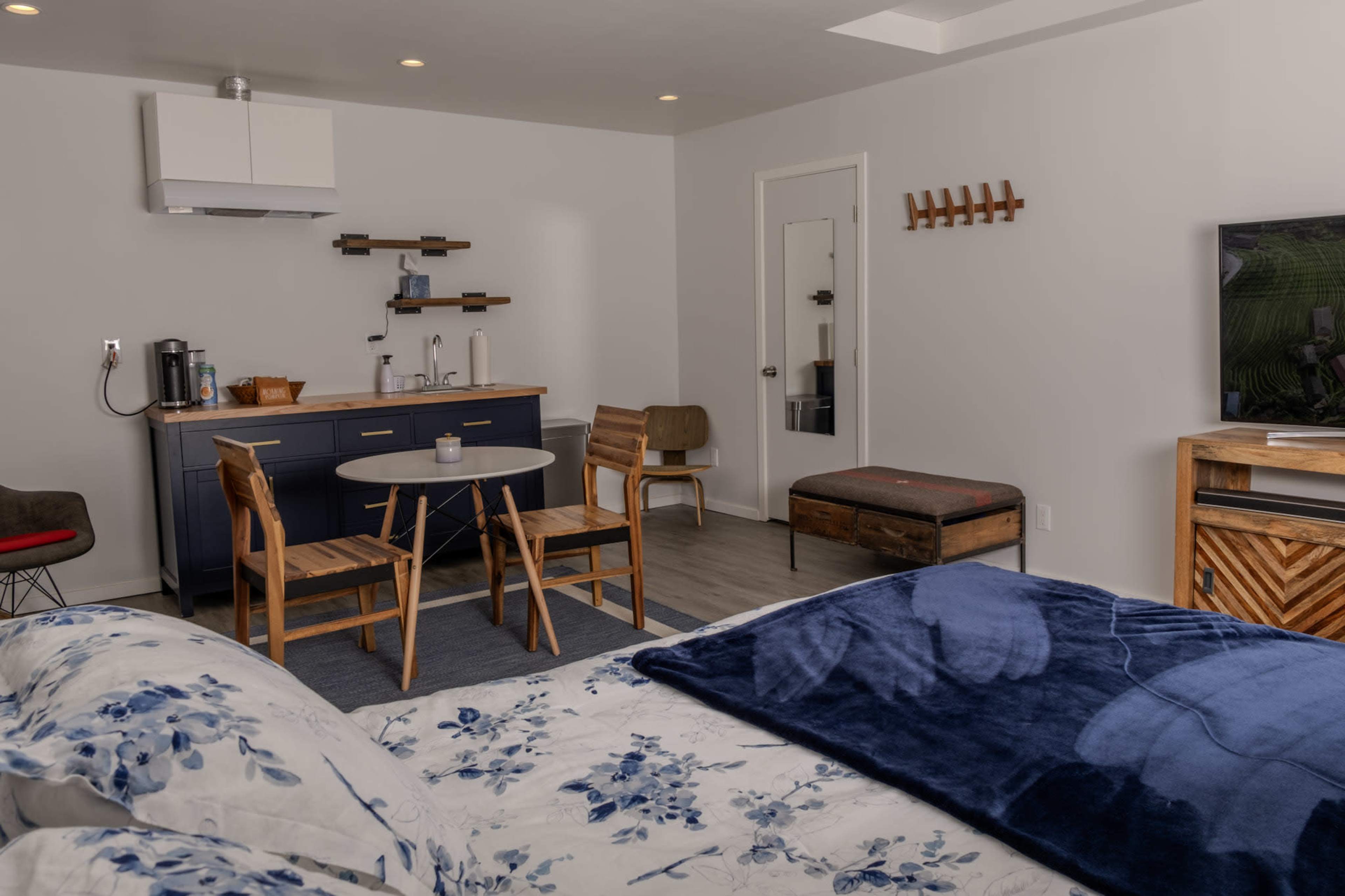 The image shows a modern living space featuring a bed, small dining table with chairs, kitchenette area, and a television.