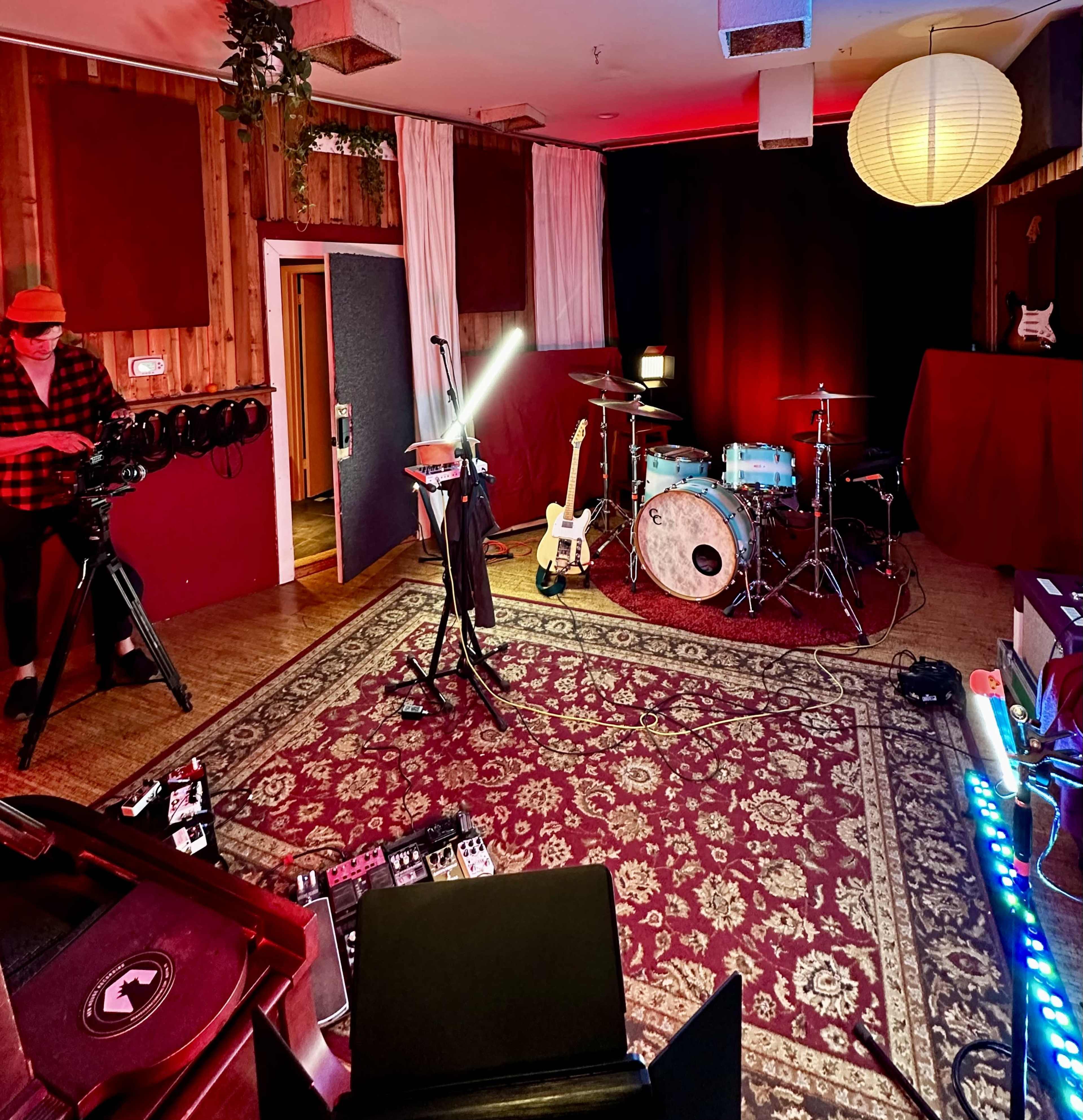 A music studio setup with instruments, including a drum kit and guitar, surrounded by various equipment and soft lighting.