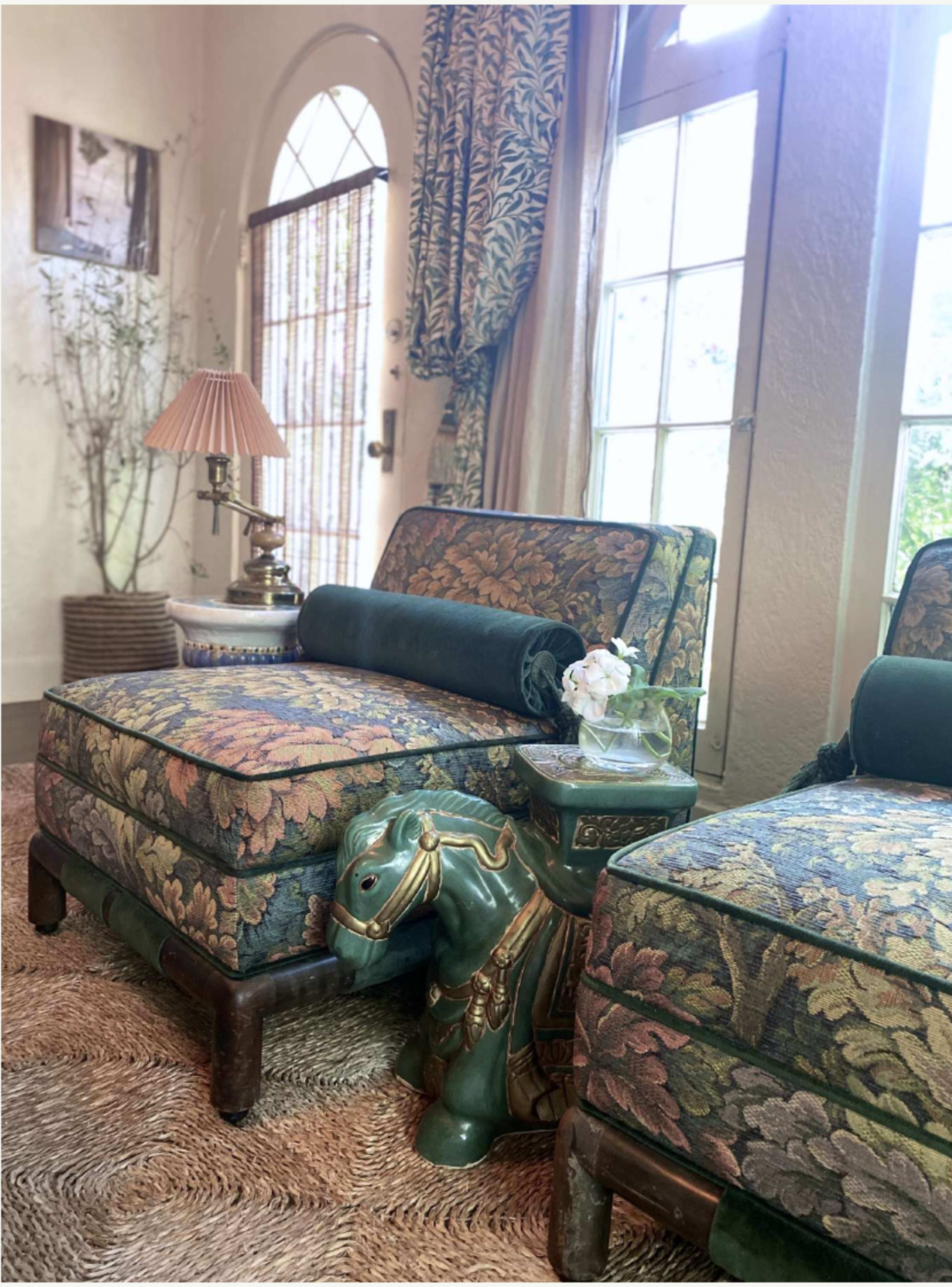 A pair of patterned chairs are positioned beside a green ceramic dog-shaped table in a sunlit room with large windows.