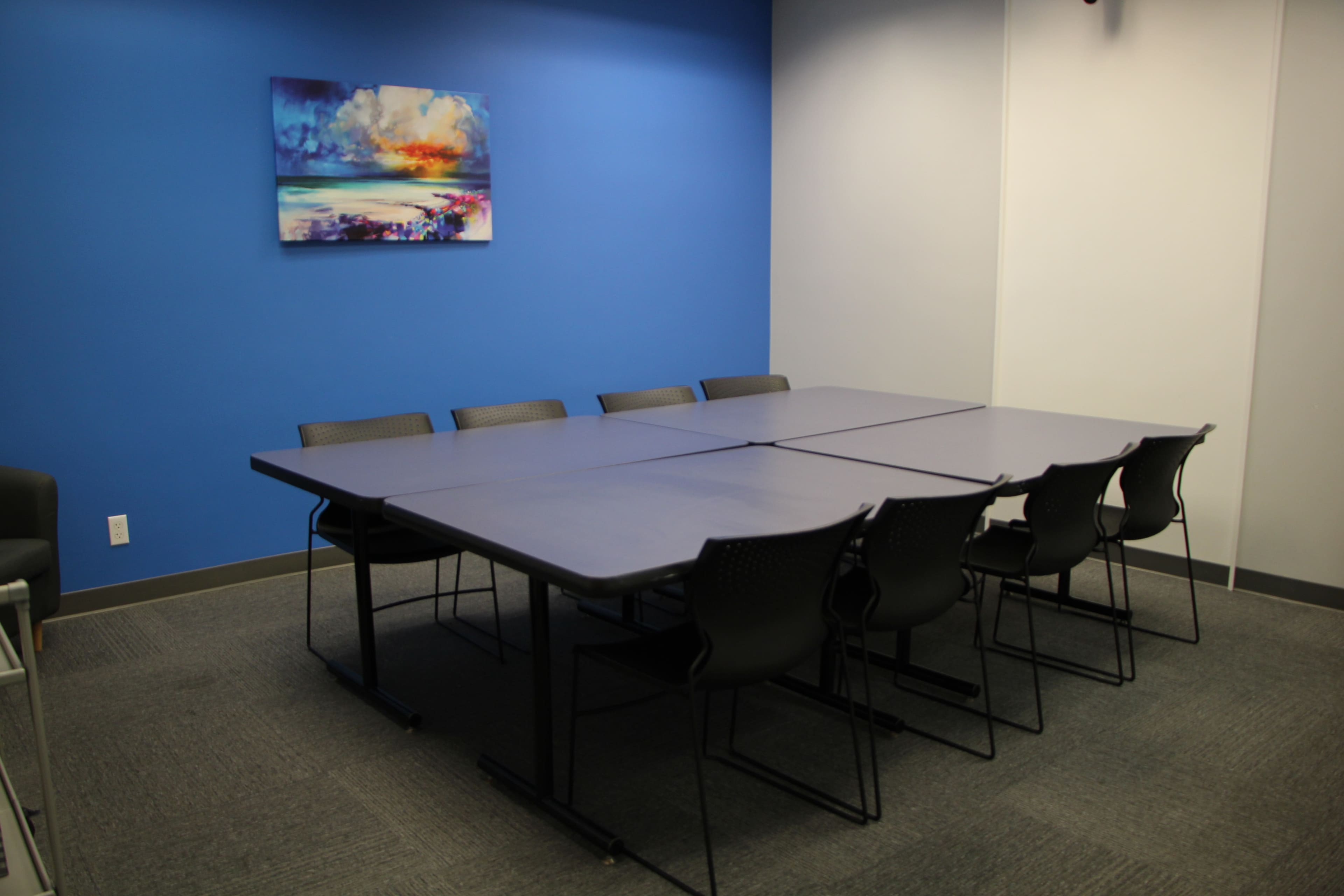 The image shows a meeting room with a large table and eight chairs, set against a blue wall and featuring a colorful abstract painting above the table.